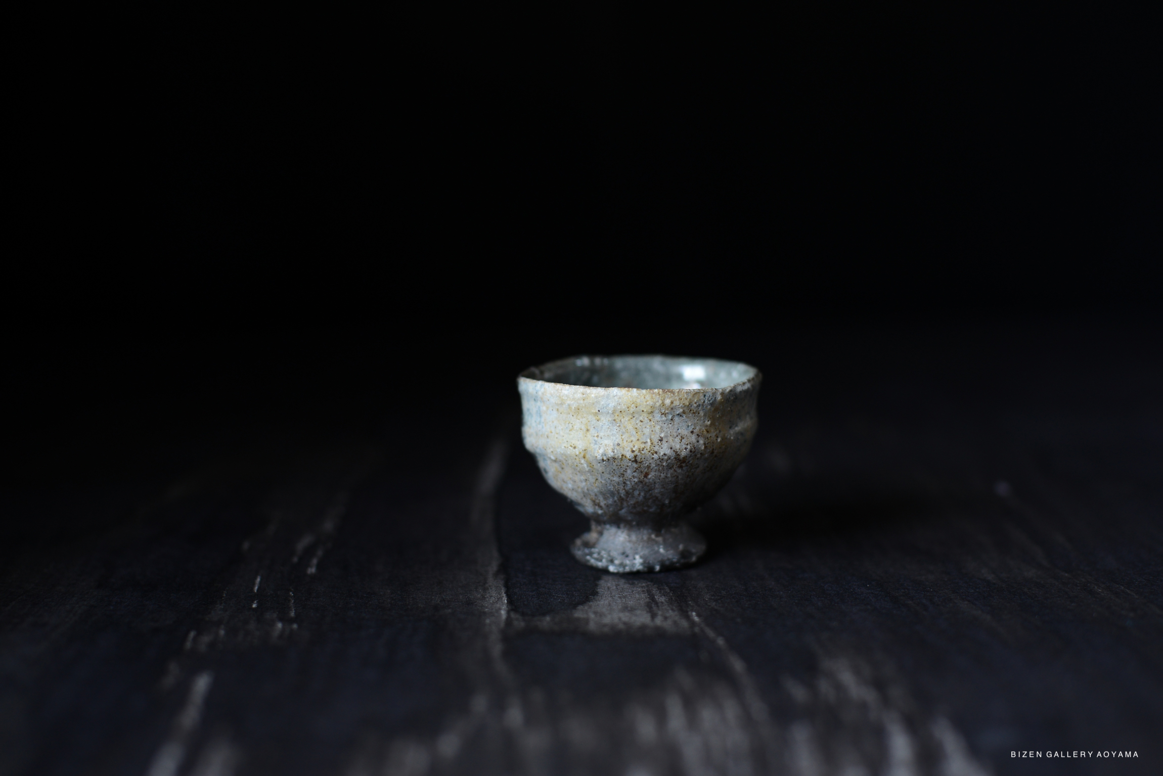 A small Iga guinomi (sake cup) with a textured surface and subtle color variations, set against a dark background.