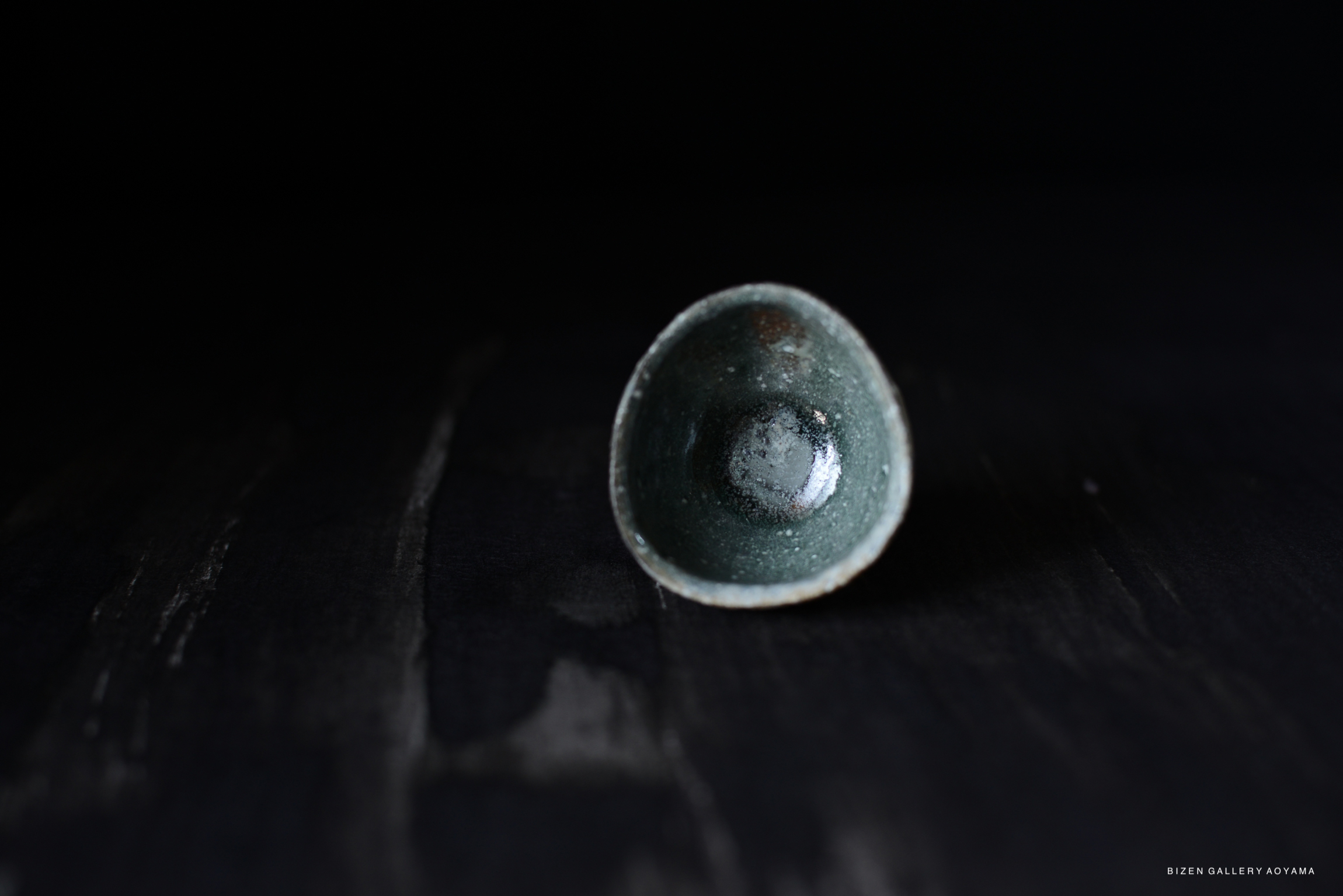 A small, round Iga pottery bowl with a glossy green glaze, placed on a dark wooden surface.