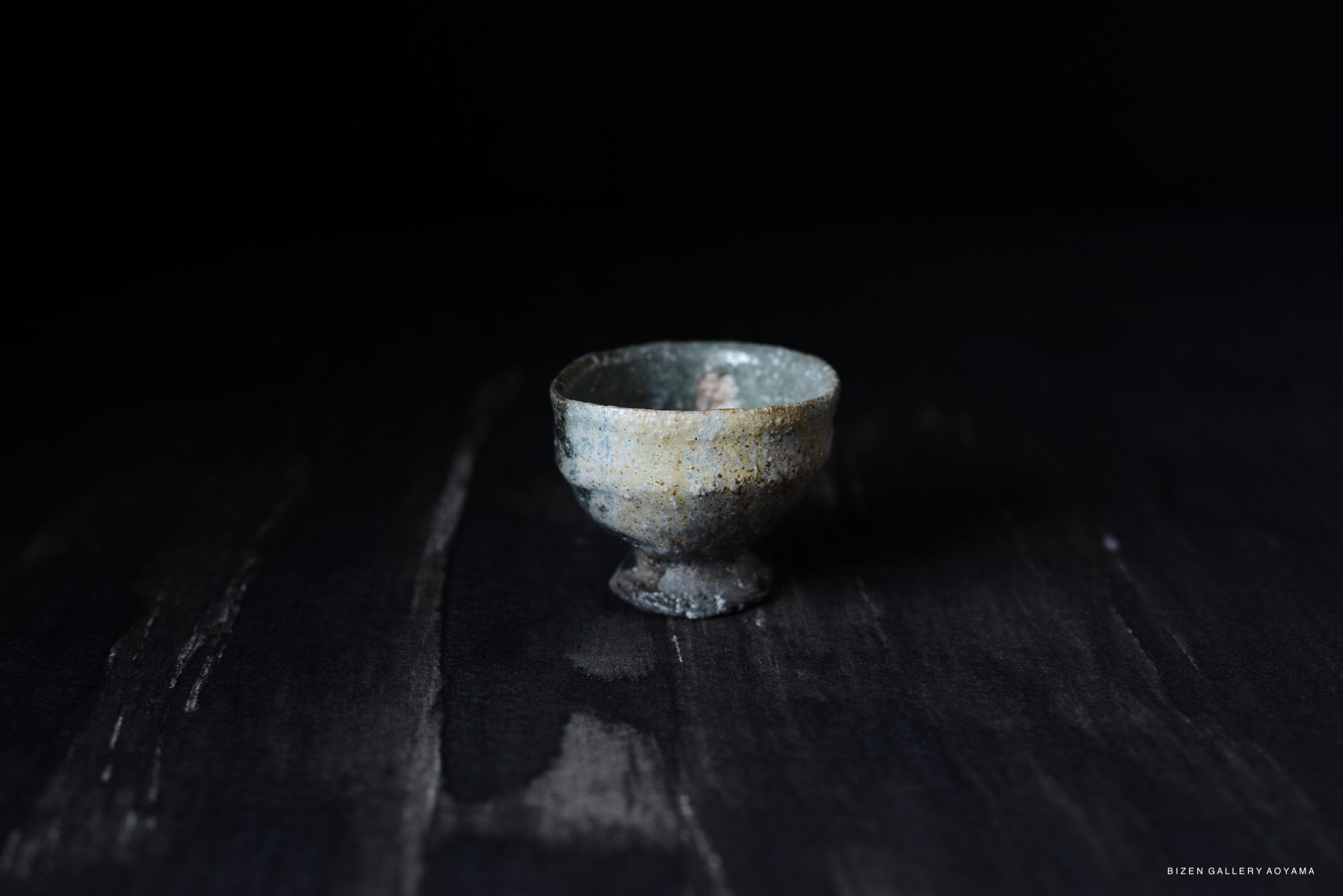 A small, rustic Iga-style guinomi (sake cup) with a textured surface and subtle color variations, set against a dark background.