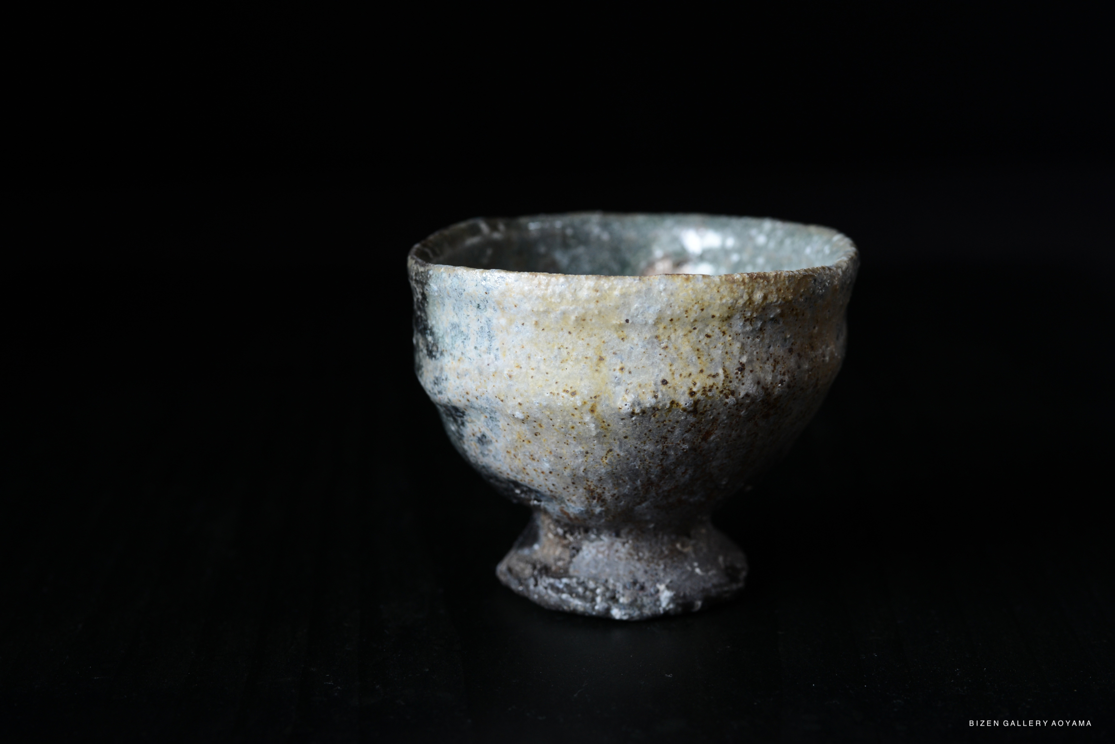 A hand-crafted Iga-style guinomi sake cup with a textured surface and a subtle gradient of colors, displayed against a dark background.