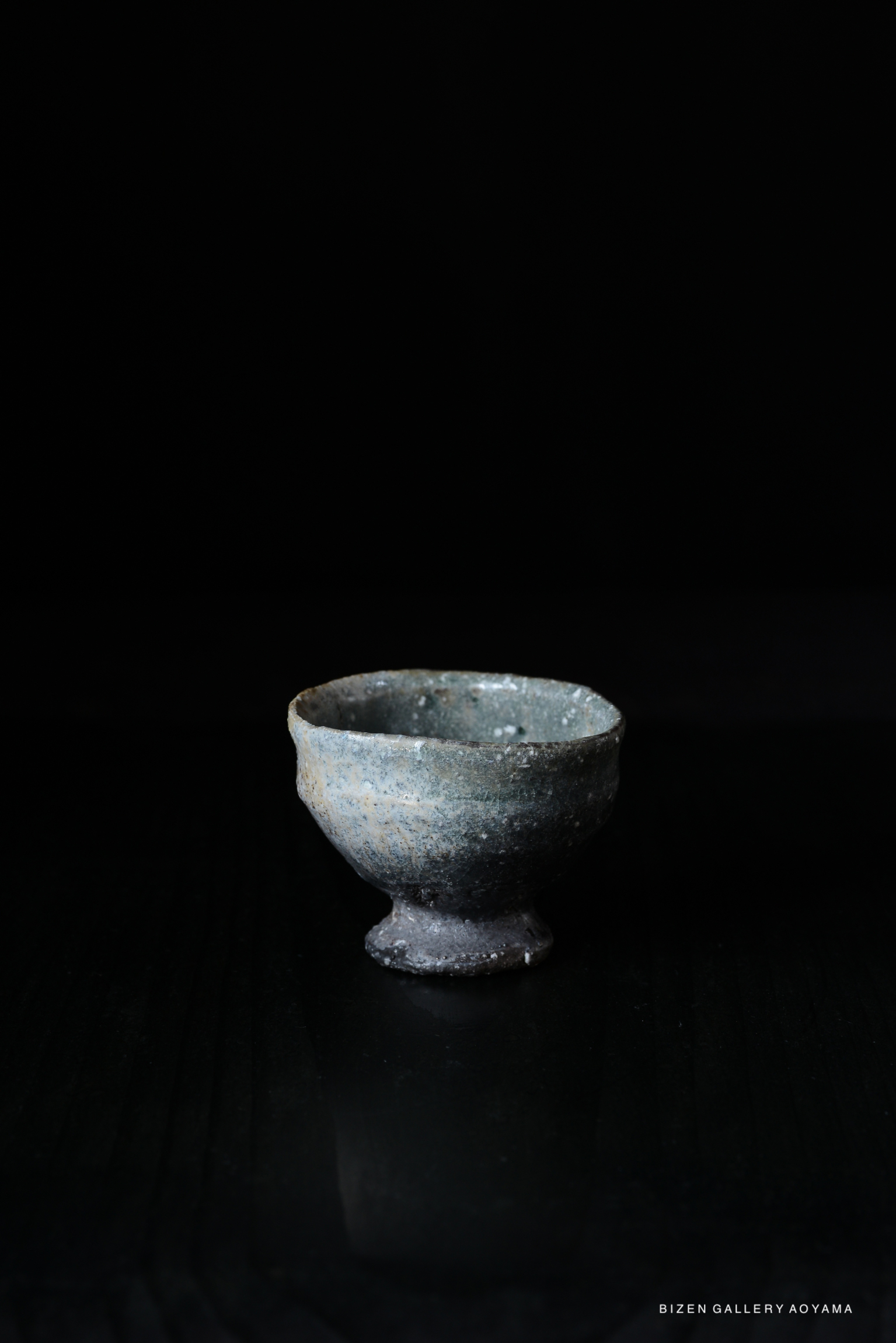 A dark, textured ceramic guinomi with a footed base, featuring a muted color palette and natural imperfections, set against a black background.