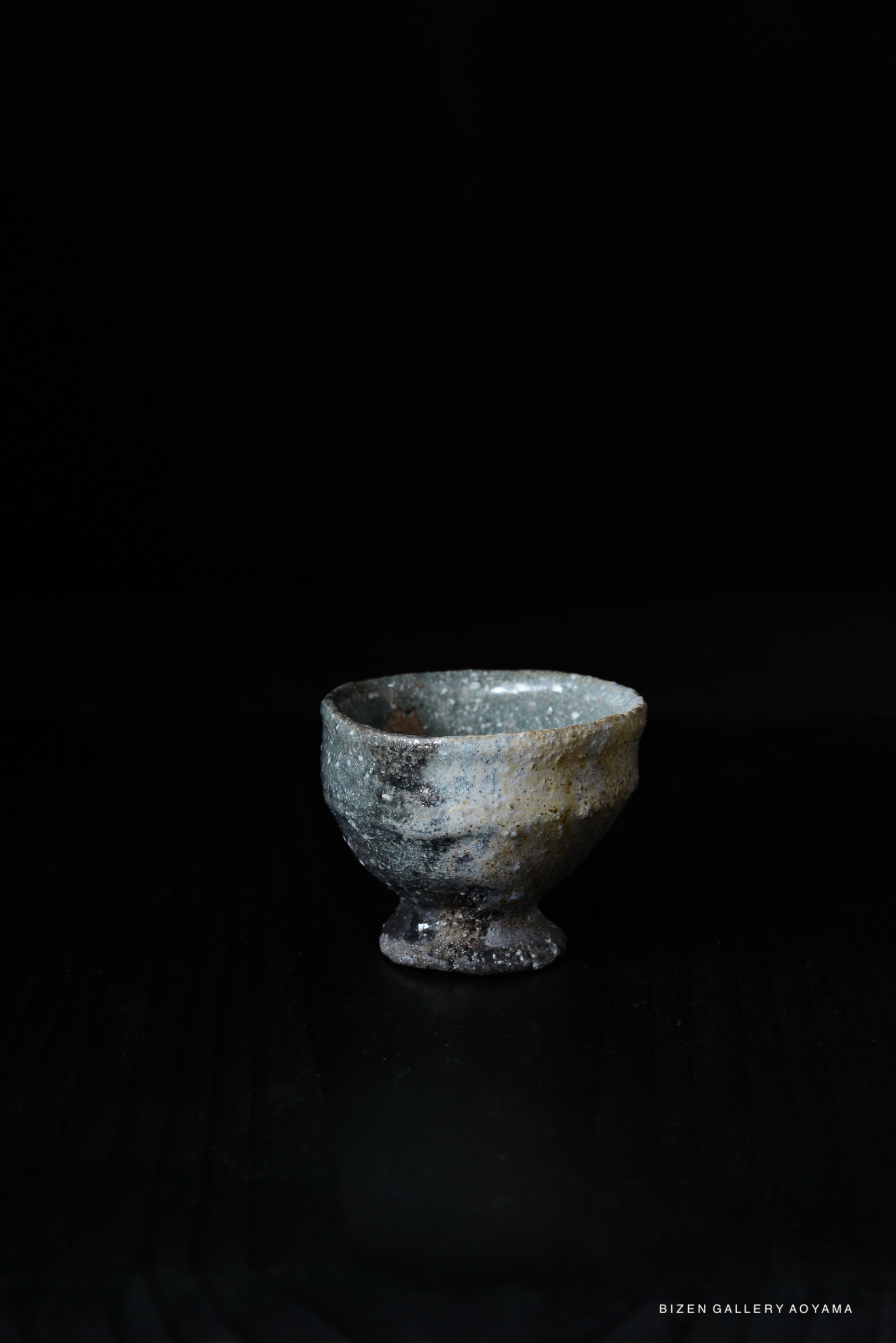 A close-up image of a traditional ceramic guinomi cup with a rustic finish, set against a dark background.