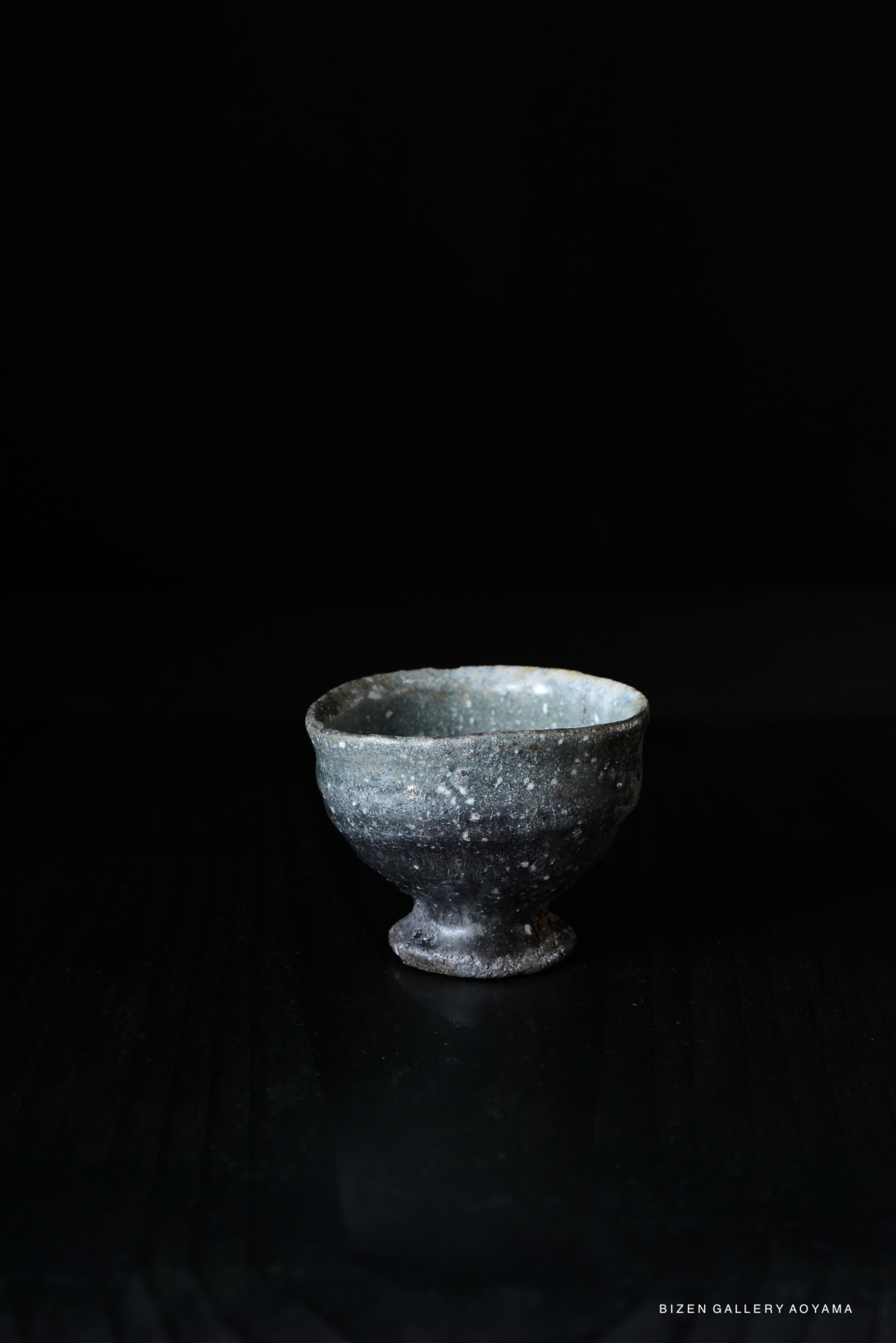 A handmade Iga wine cup with a rustic texture and dark coloration, displayed on a dark surface.