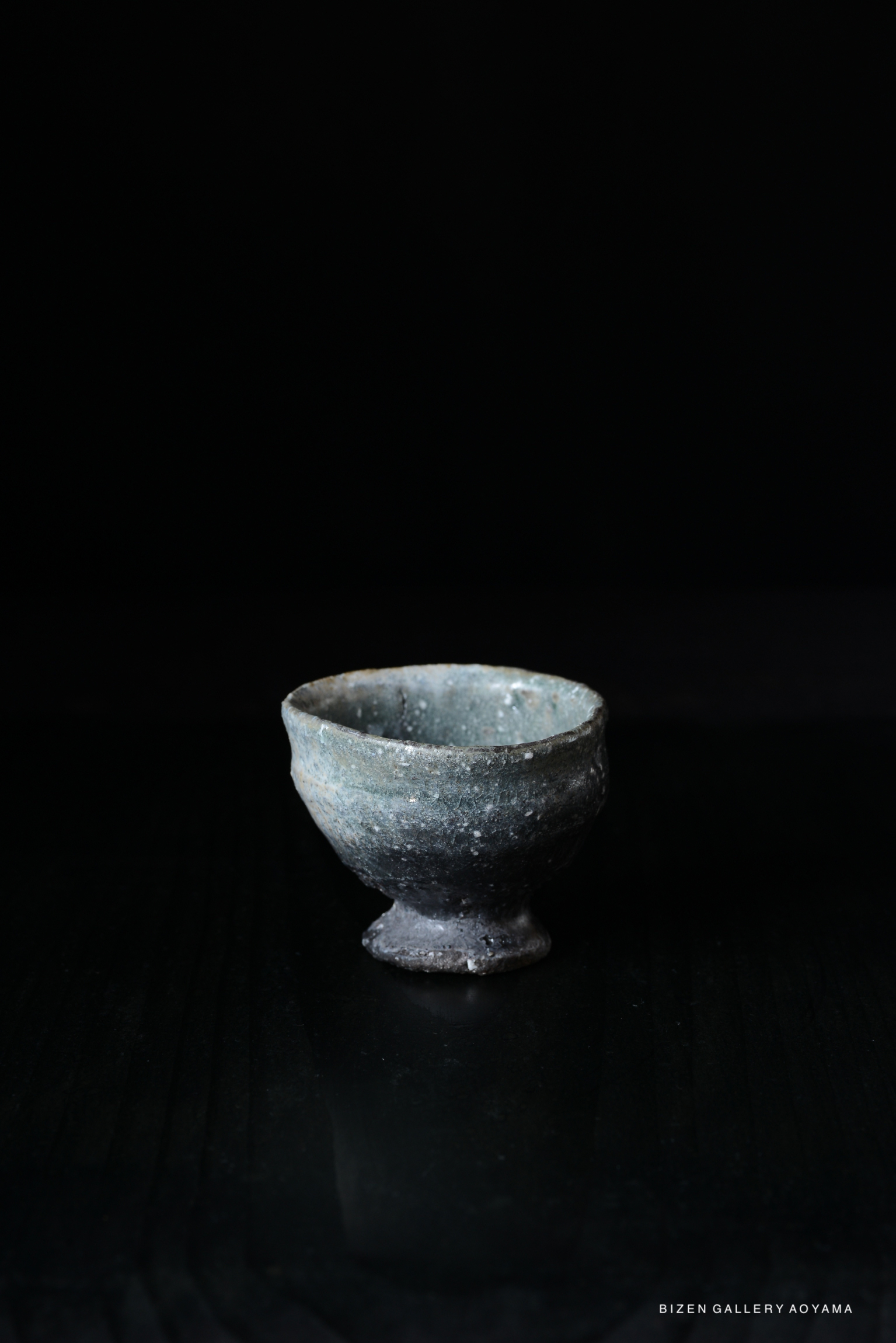 A close-up of a unique, rustic Iga-style guinomi (sake cup) with a footed base, featuring a textured surface and earthy tones, set against a dark background.
