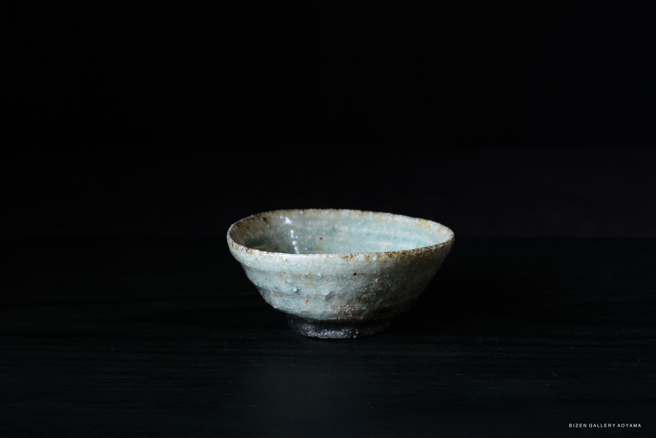 A small, rustic ceramic bowl with a light green glaze and visible texture, set against a dark background.