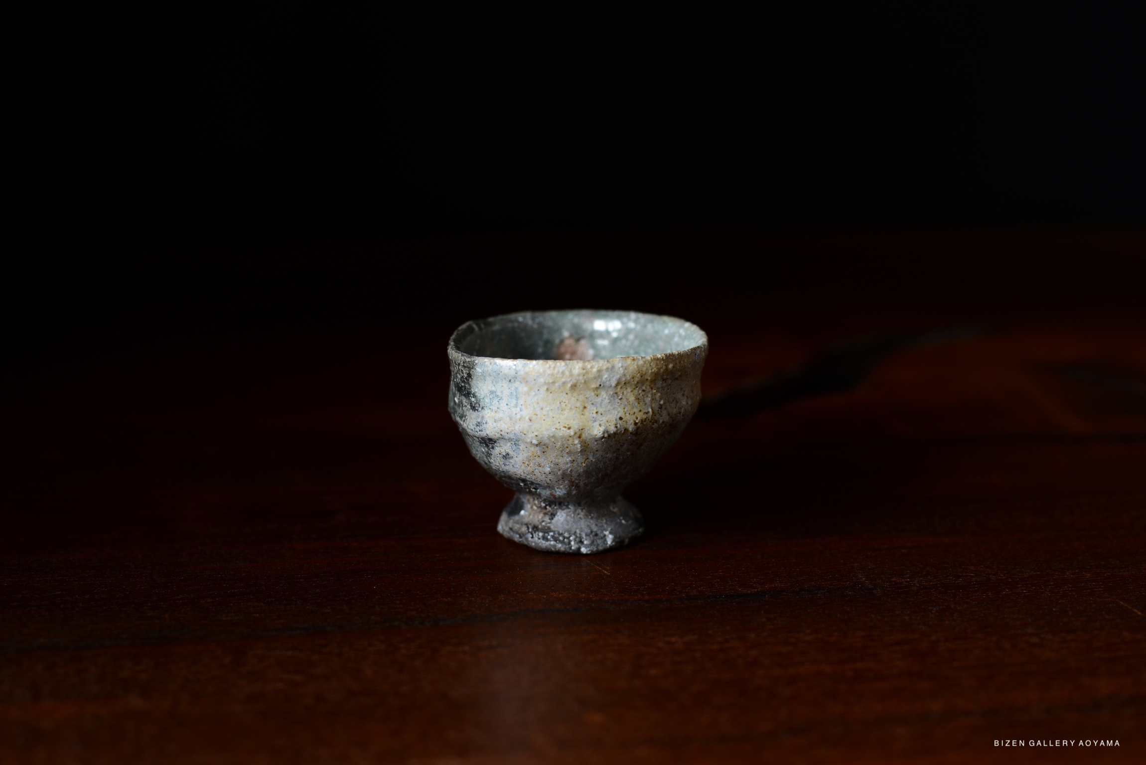 A small, handcrafted Iga cup featuring a rustic, speckled glaze, placed on a dark wooden surface.