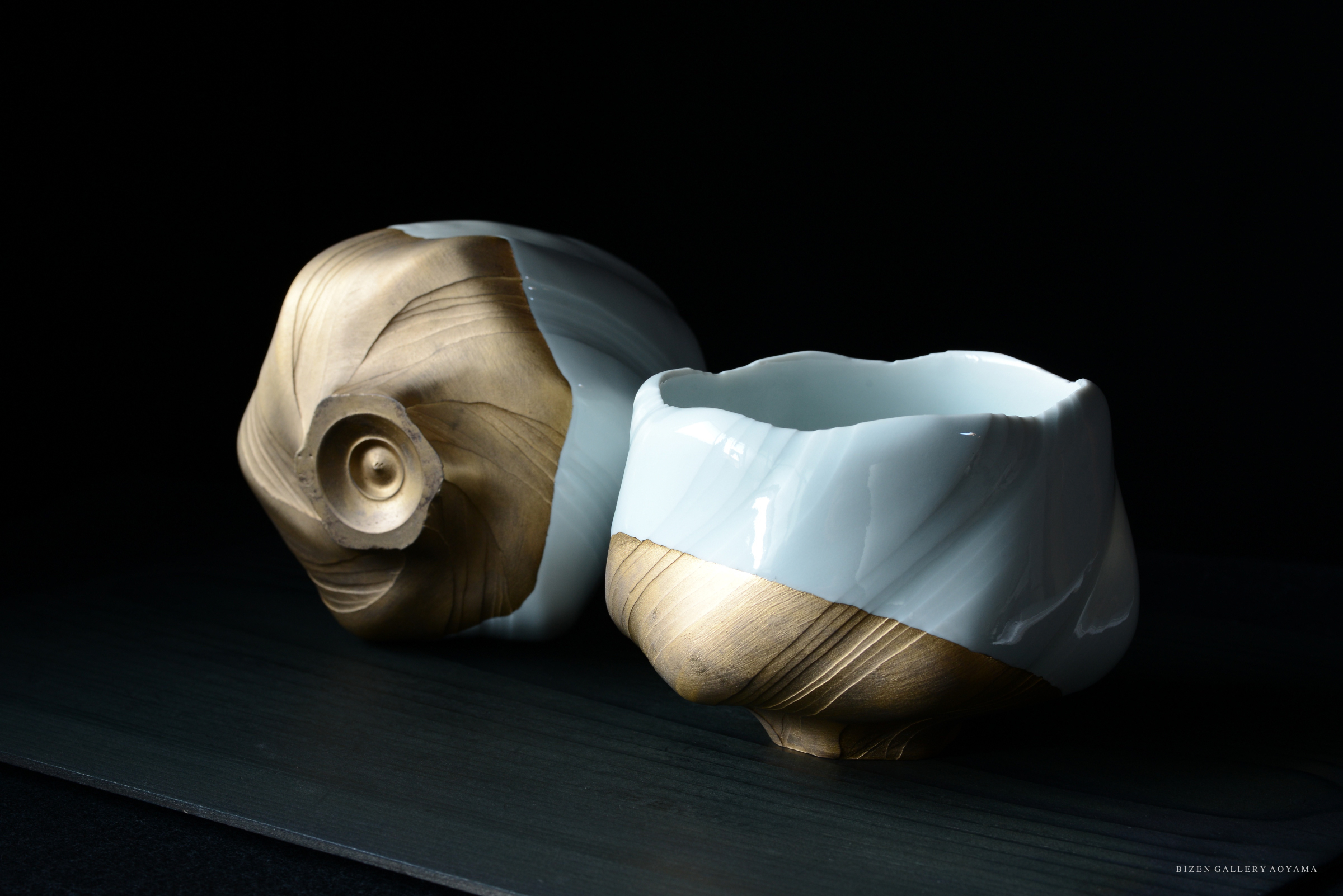 A close-up view of two elegant ceramic pieces, featuring a shiny gold and white finish, resembling the shape of seashells or natural forms, against a dark background.