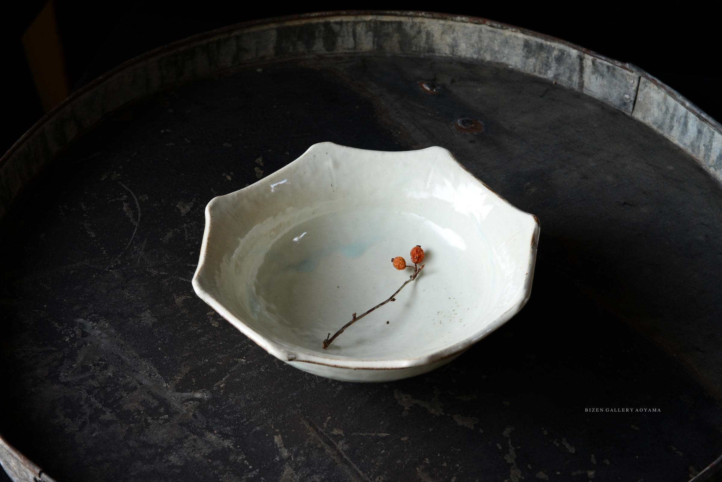 A delicate, white ceramic bowl with an octagonal shape, resting on a dark, textured surface, featuring a twig with red berries inside.