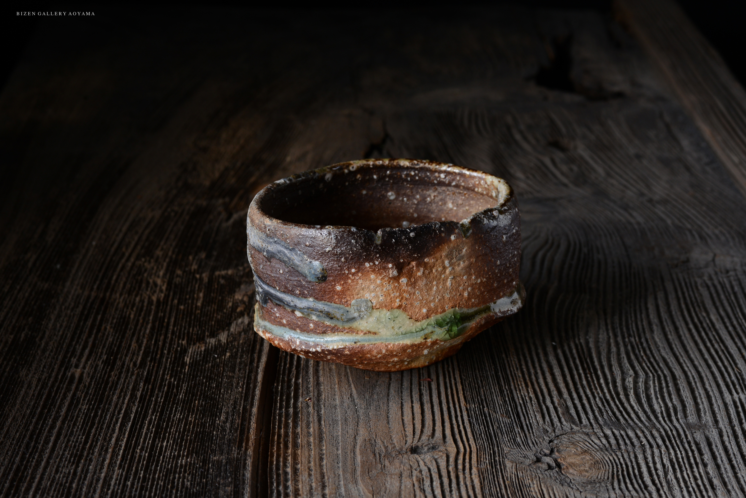 陶芸の器が暗い木のテーブルの上に置かれています。器の表面には独特な釉薬の模様と質感があり、自然な色合いが特徴です。