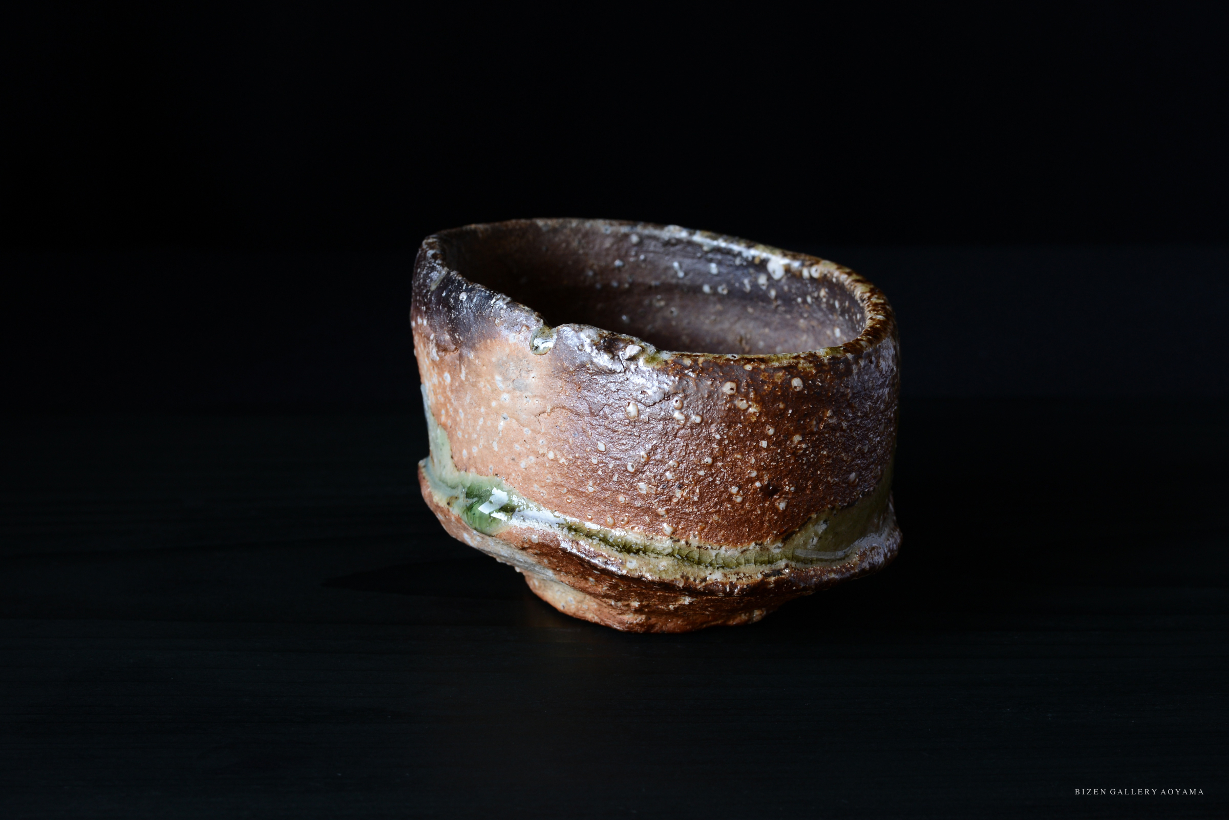 黒い背景の上に置かれた、手作りの陶器の鉢。表面はざらざらしており、色合いは茶色と緑が混ざった風合い。