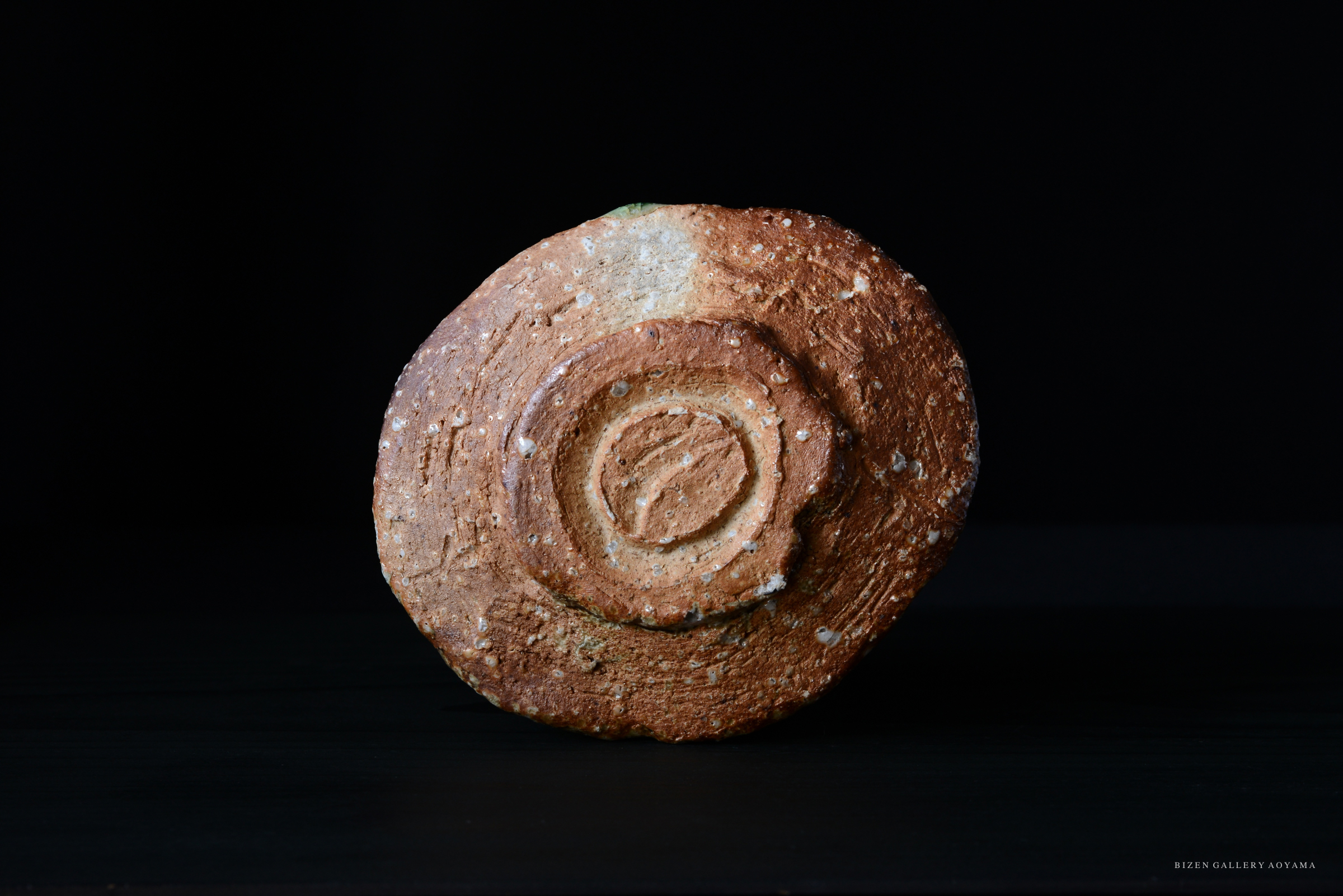 黒背景の上に置かれた、円形の陶器のオブジェ。表面はざらざらしており、茶色と緑色の部分が見られる。中央には円形の模様が彫られている。