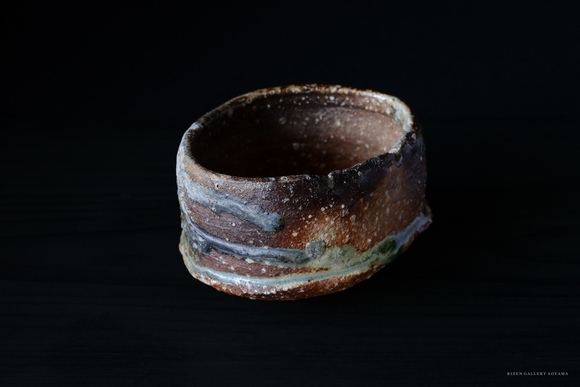 黒い背景に置かれた、土の質感を持つ陶器の器。表面には複数の色の釉薬の線が見られる。