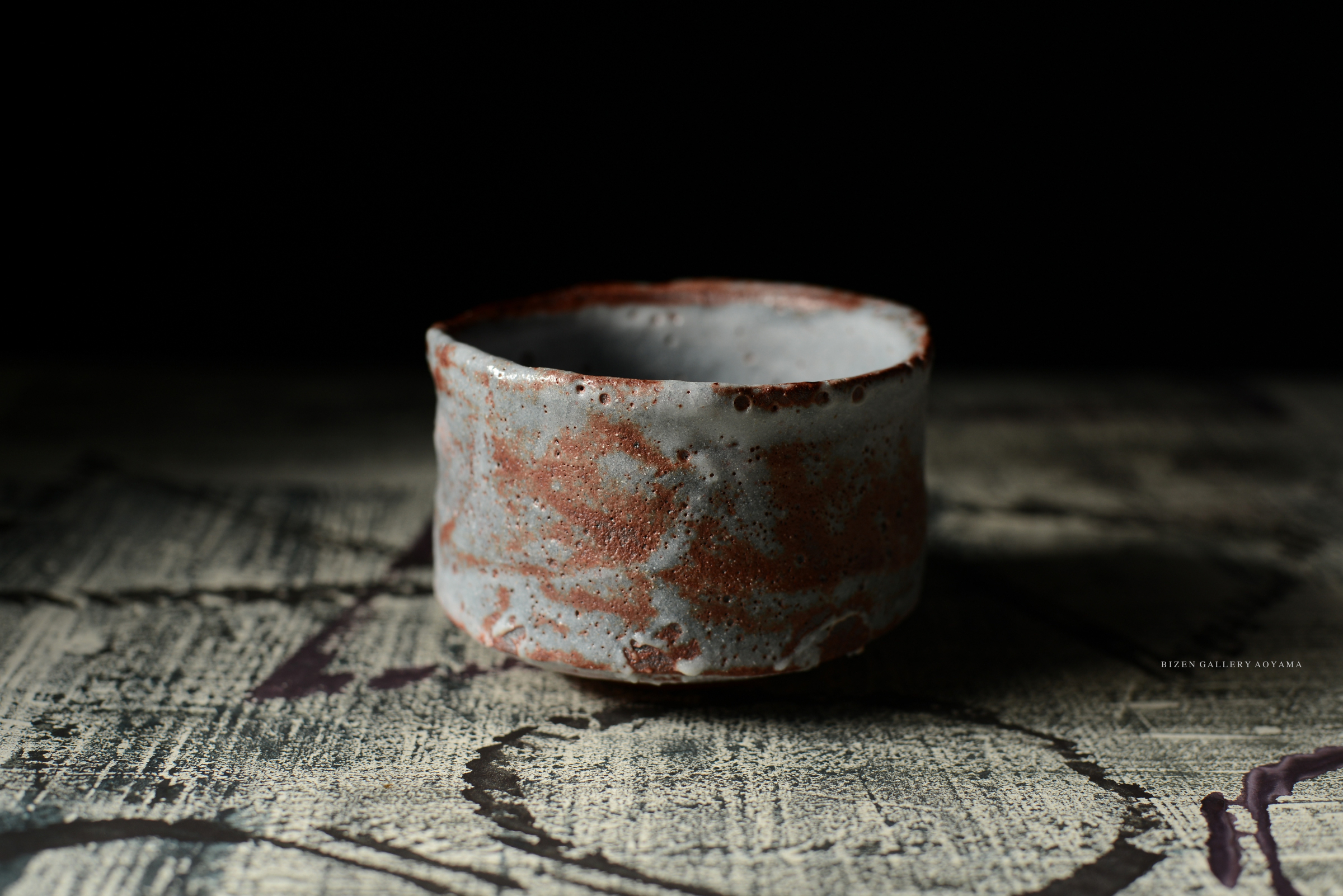 A close-up of a rustic ceramic tea bowl with a textured grey and reddish-brown surface, placed on a patterned table.