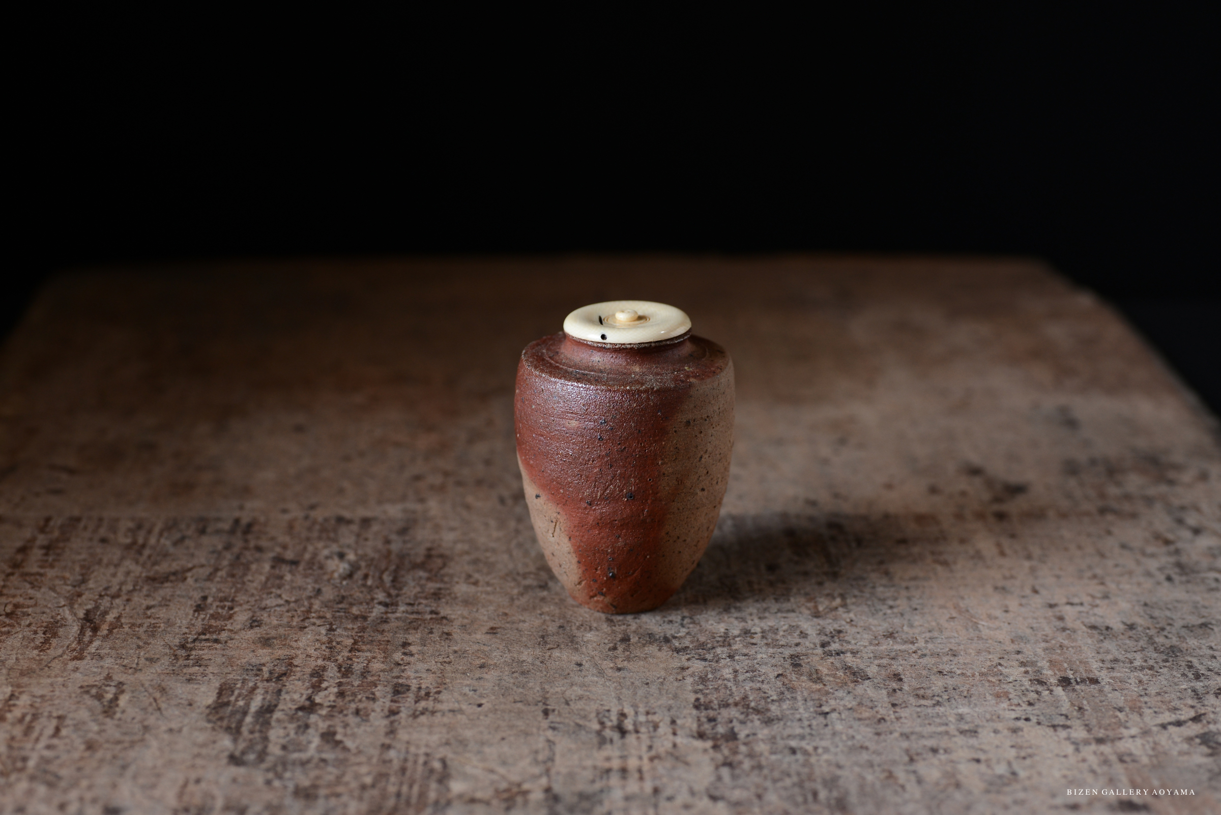 A small pottery container with a textured brown surface and a light-colored lid, sitting on a rustic wooden surface.