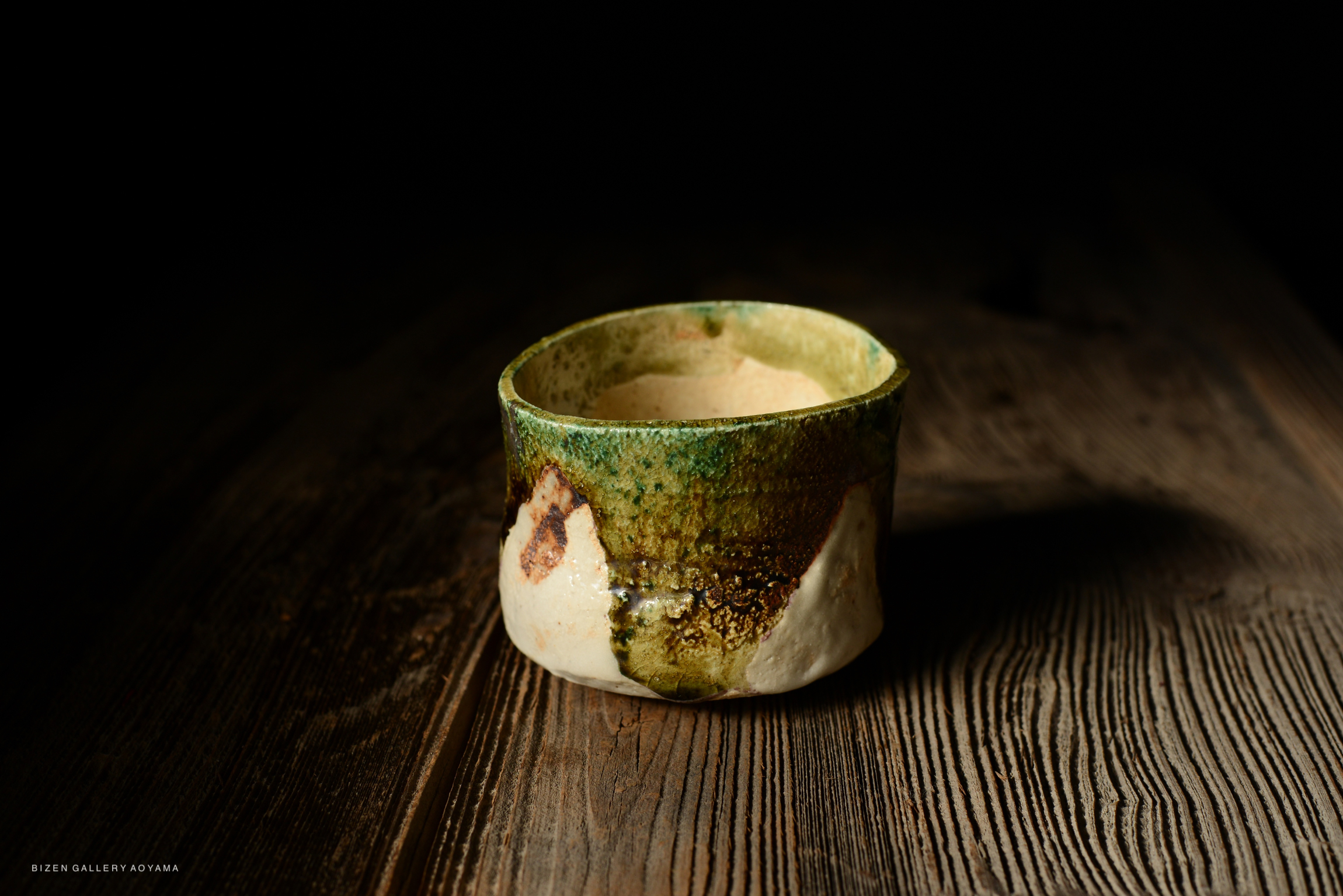 A ceramic cup with a green and brown glazed surface, resting on a wooden table.