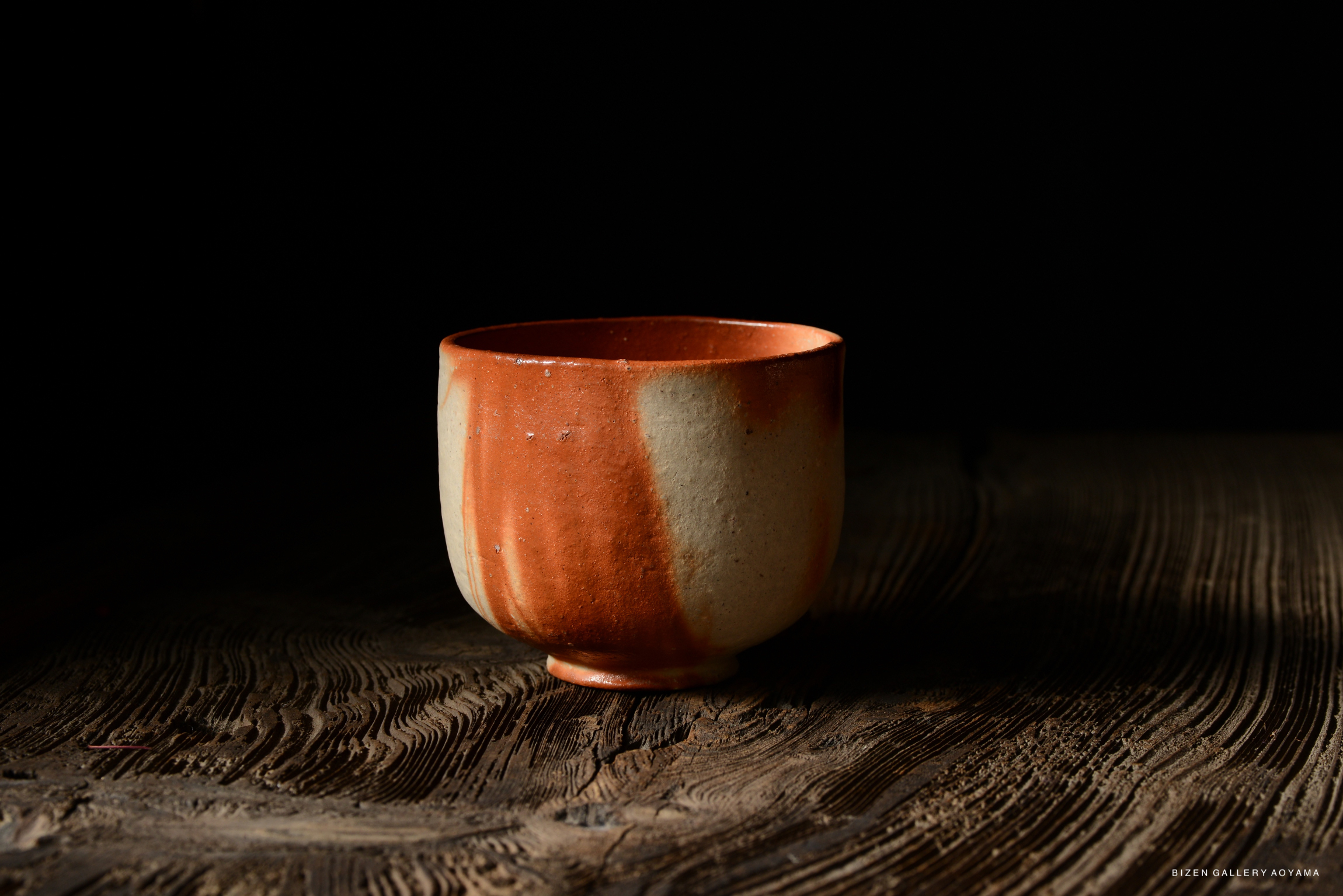 A Bizen Hidasuki Chawan, showcasing a moist scarlet color and a textured clay surface, placed on a wooden table.