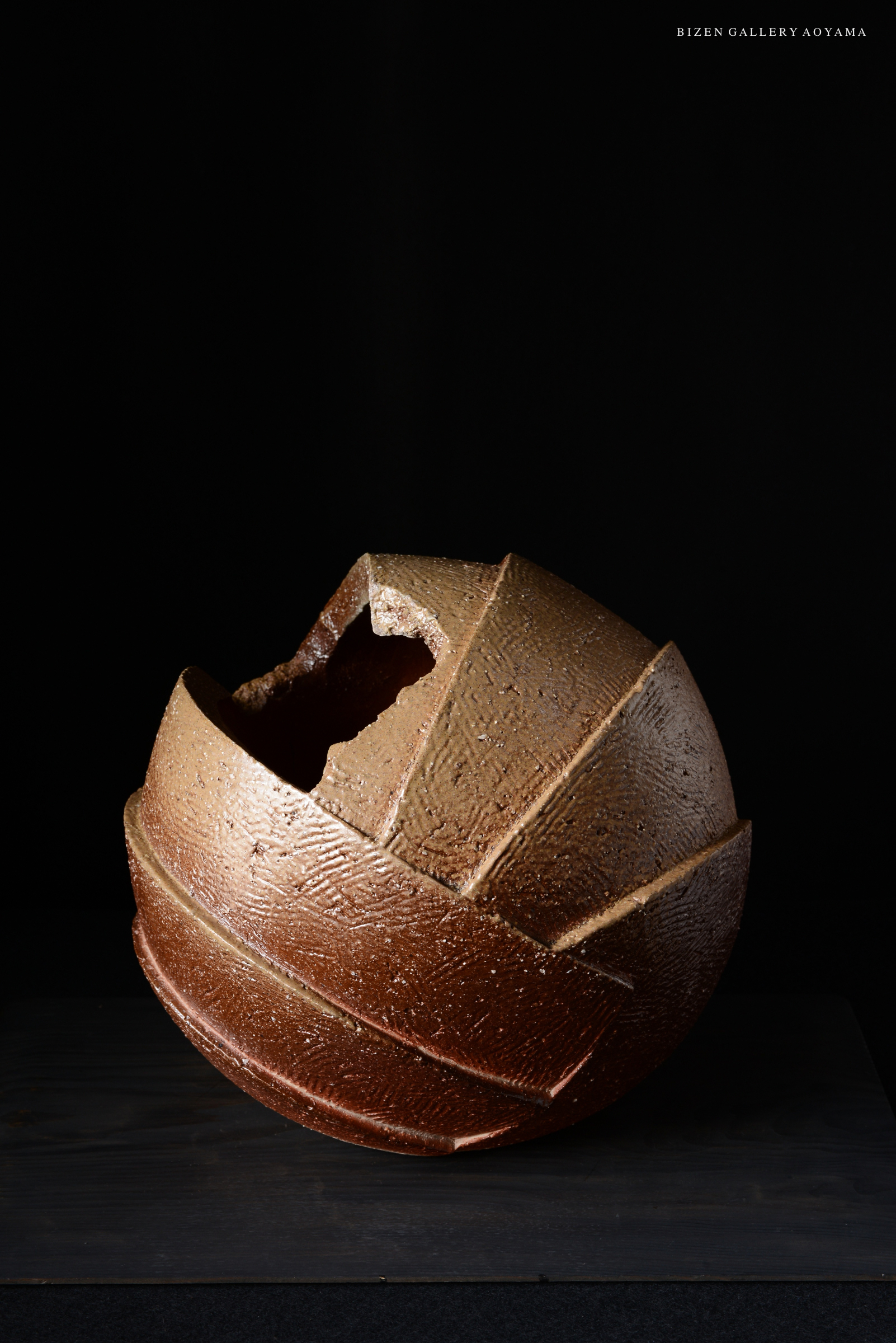 A Bizen Shinogi Tsubo, a traditional Japanese pottery piece, featuring a unique round shape with a textured surface and a broken section revealing its inner structure. The pot is displayed on a dark background.