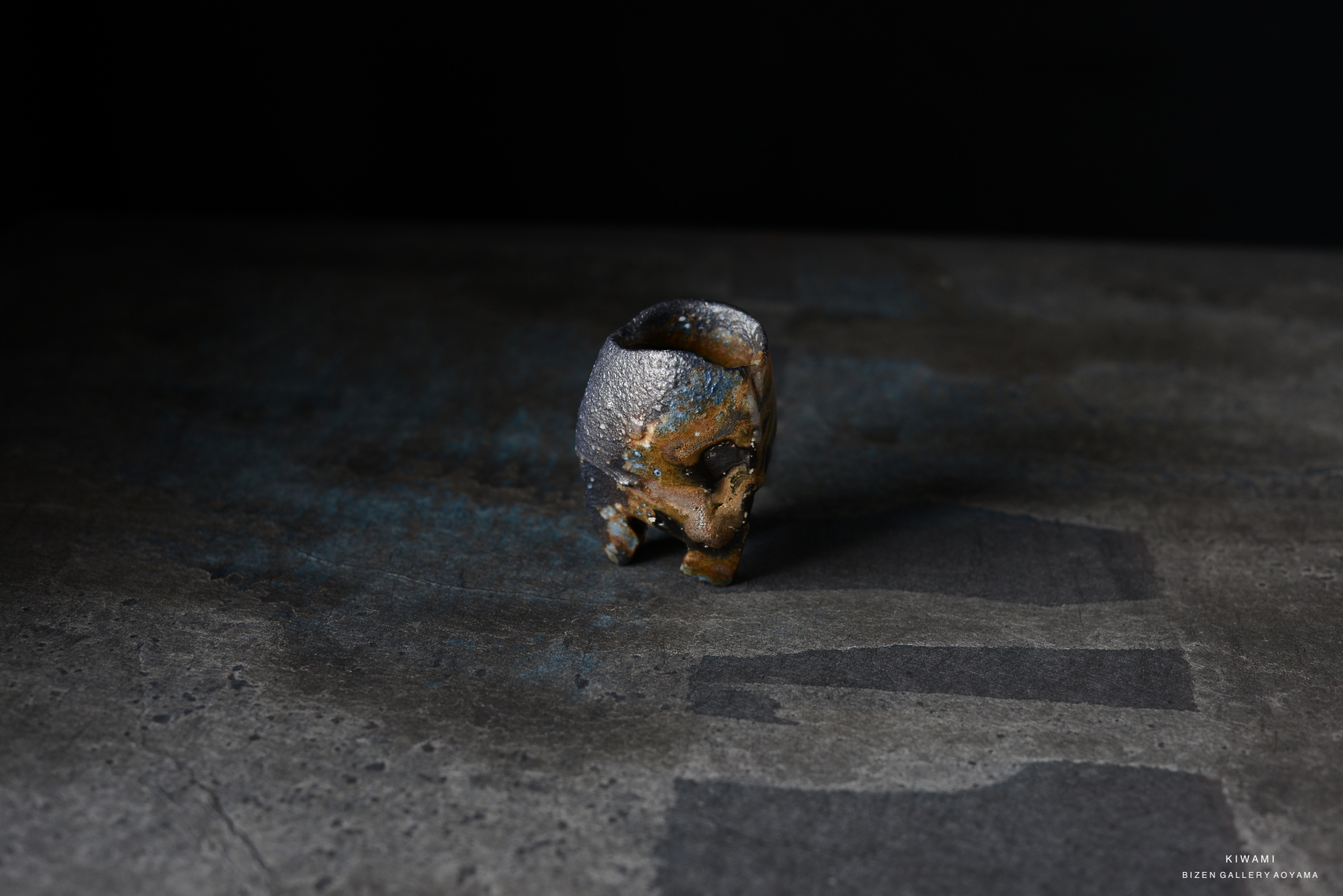 A close-up of a unique ceramic sake cup (kuro guinomi) with a textured, dark finish and earthy colors, set against a dark background.