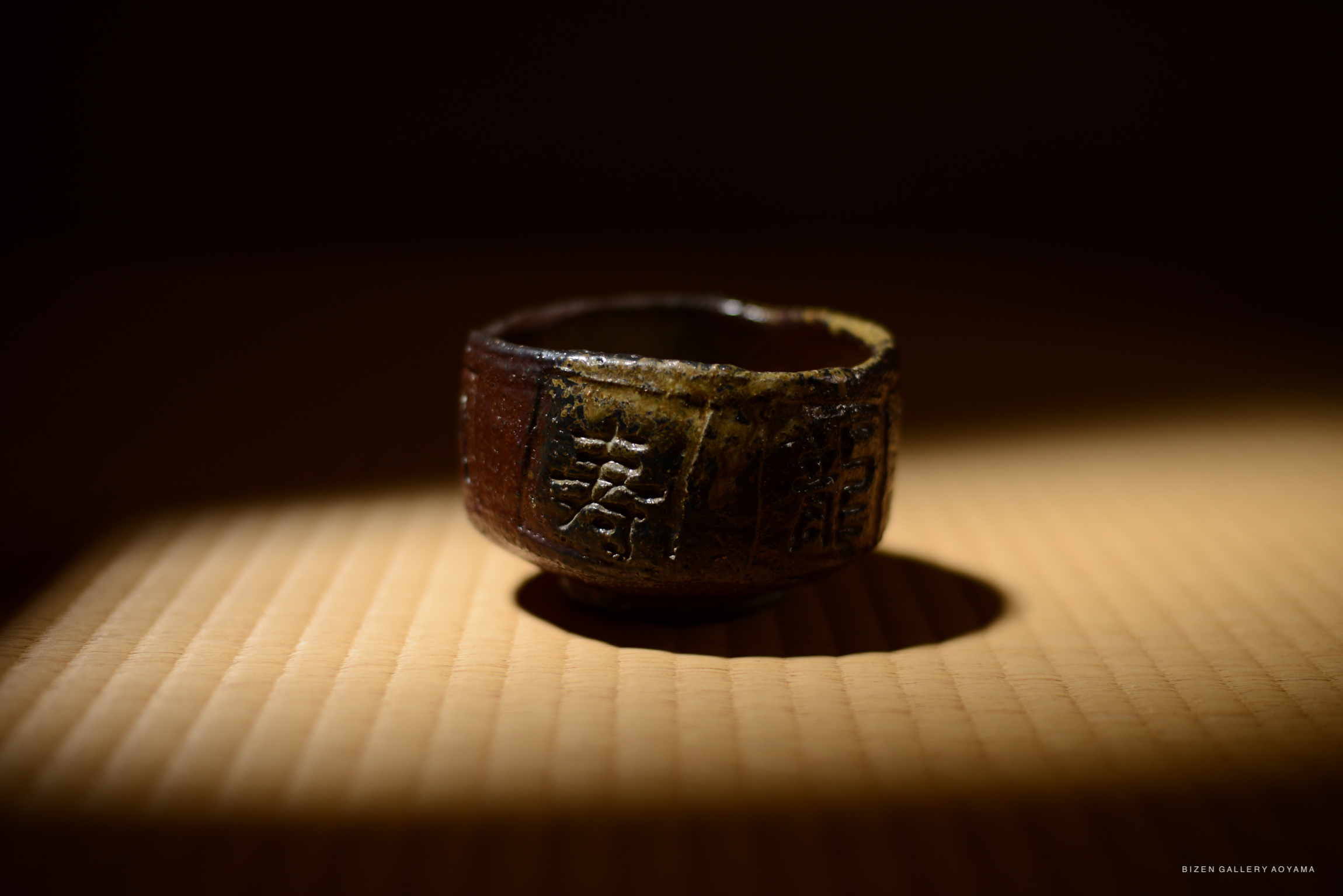 A close-up image of a Bizen-style tea bowl with auspicious characters engraved, showcasing a deep reddish-brown surface with a melted chocolate texture, illuminated by soft light.