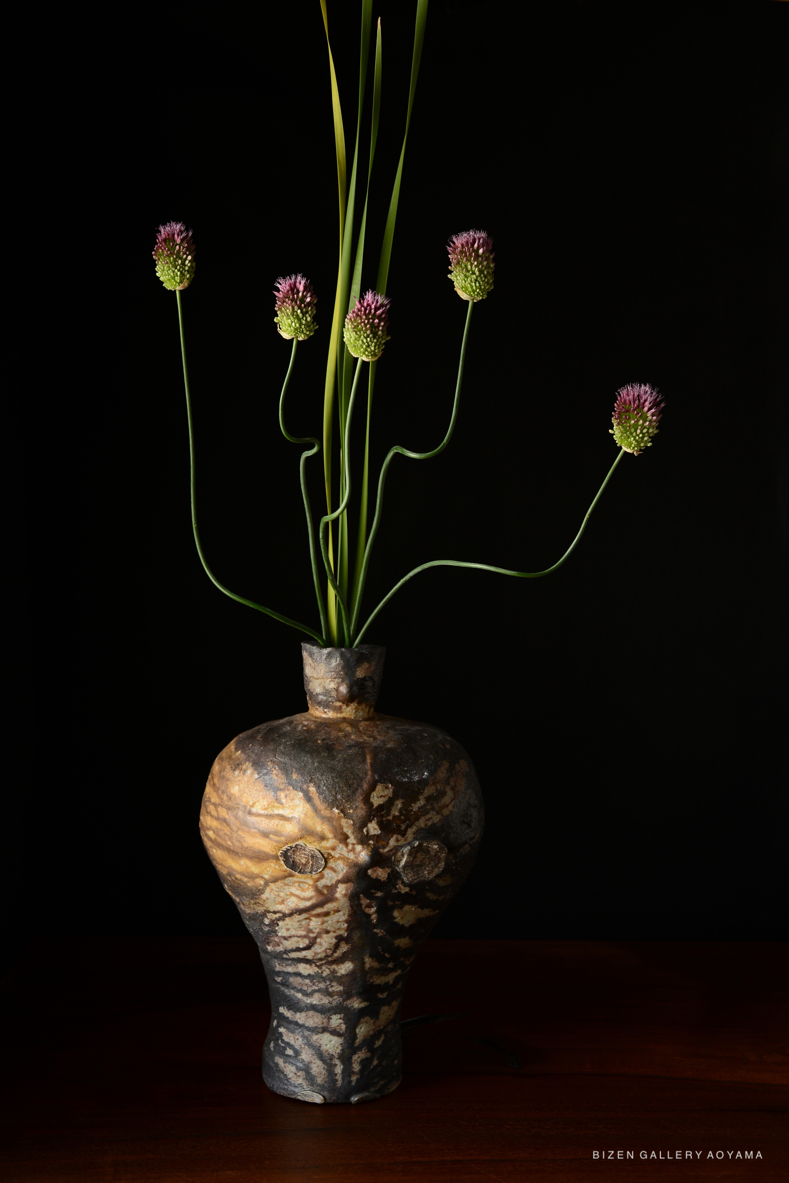 A decorative Bizen vase with a unique, textured surface, displaying tall green stems and pinkish flower buds against a black background.