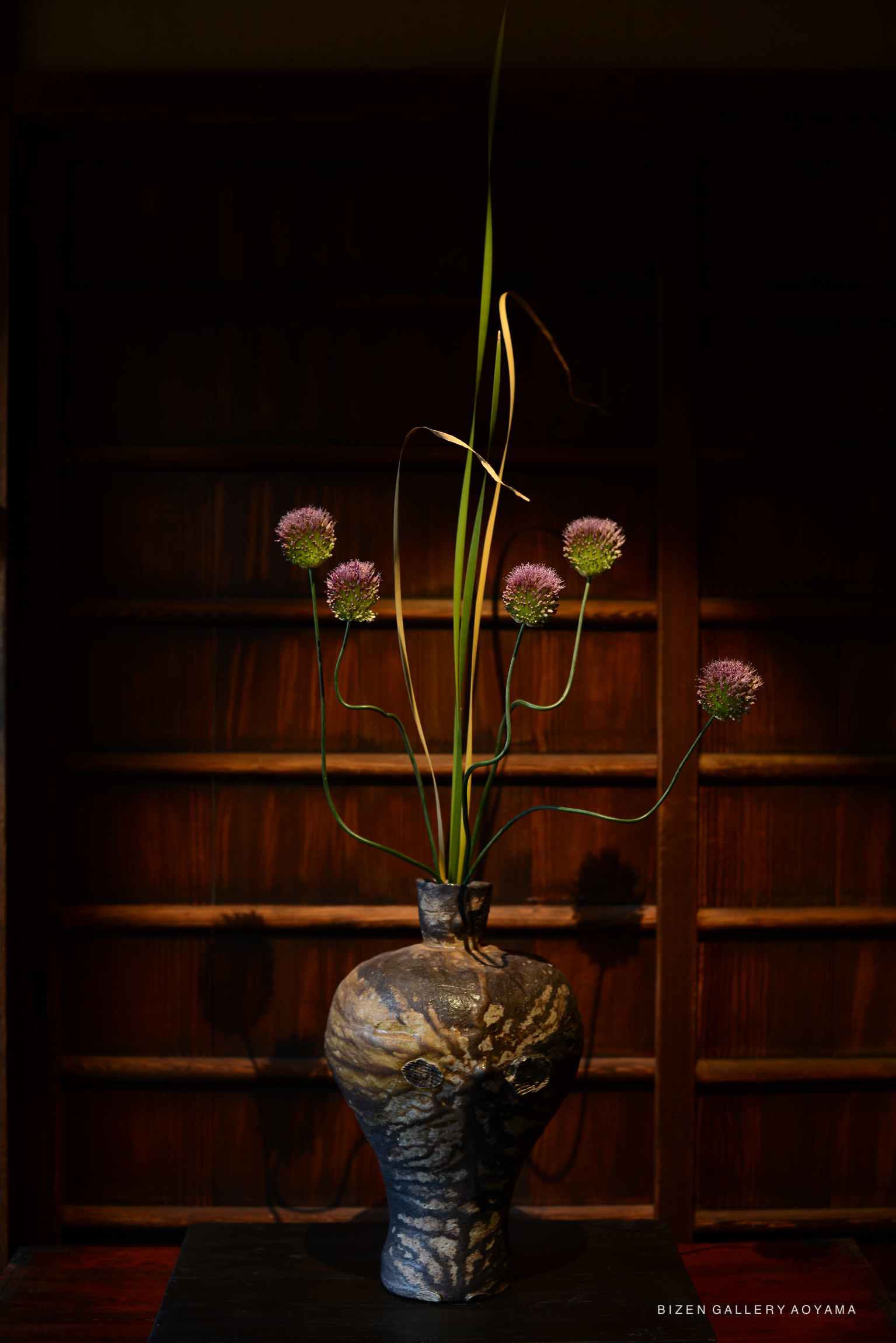 An artistic Bizen pottery vase by Sei Hoshino, featuring an elongated neck and abstract design, holding green shoots and purple flowers, set against a wooden background.