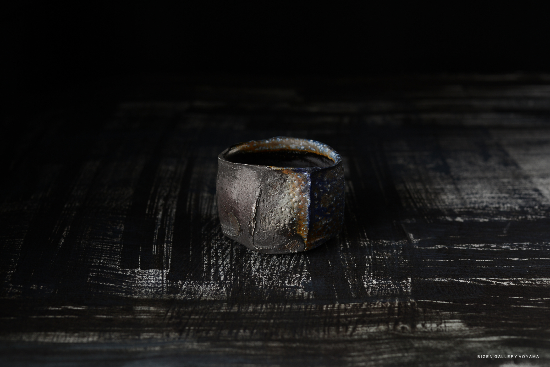 A Bizen Kofukuwan, a small ceramic bowl with a textured dark surface and subtle color variations, displayed on a dark textured background.