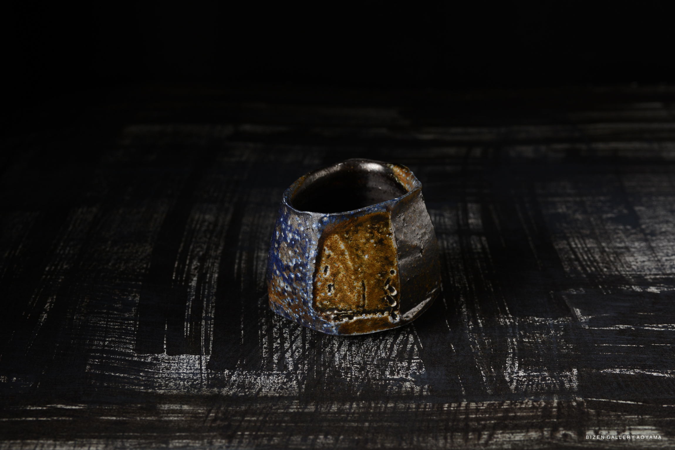 A close-up view of a Bizen pottery Kofuku Wan (小服碗), featuring earthy tones with blue and brown glazes, on a textured black surface.