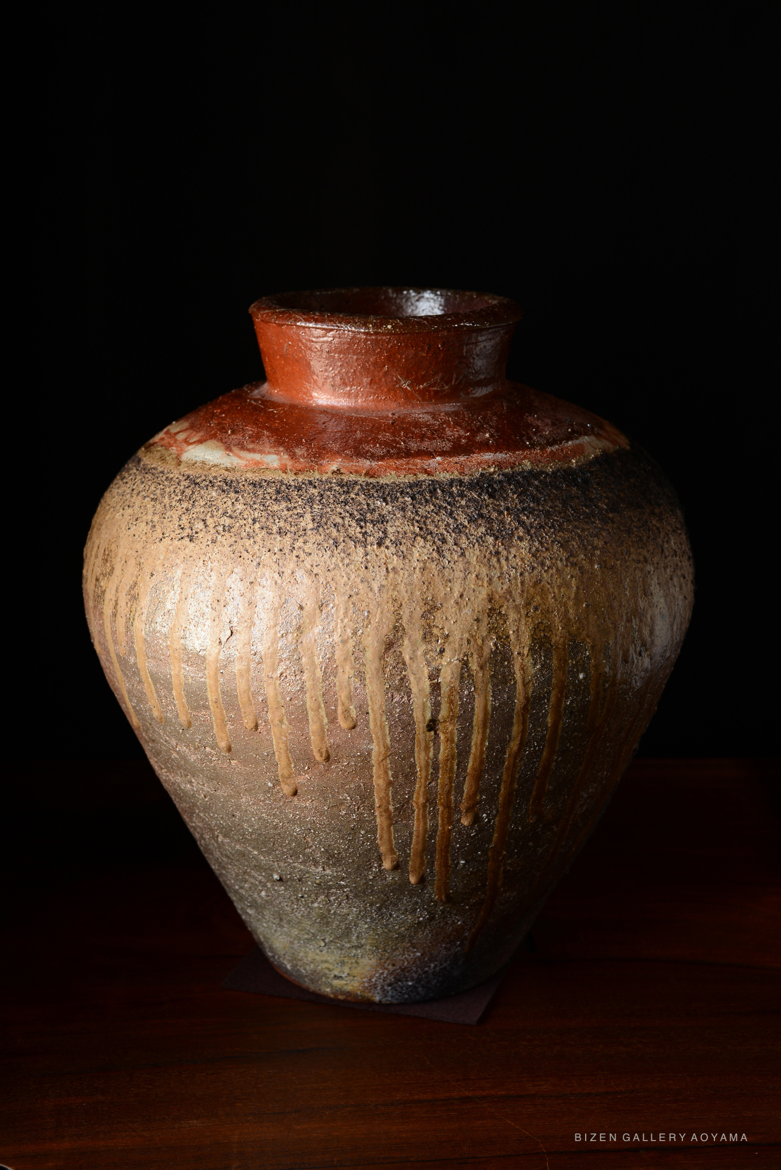 A traditional Bizen Tsubo pottery jar with a round body and narrow neck, featuring earthy tones and dripped glaze effects, set against a dark background.