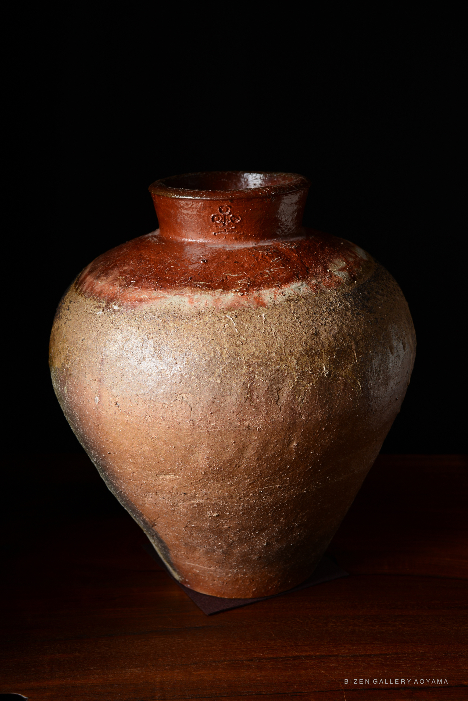 A traditional Bizen tsubo, a Japanese ceramic storage jar, featuring a rounded body and a narrow neck, displayed against a dark background.