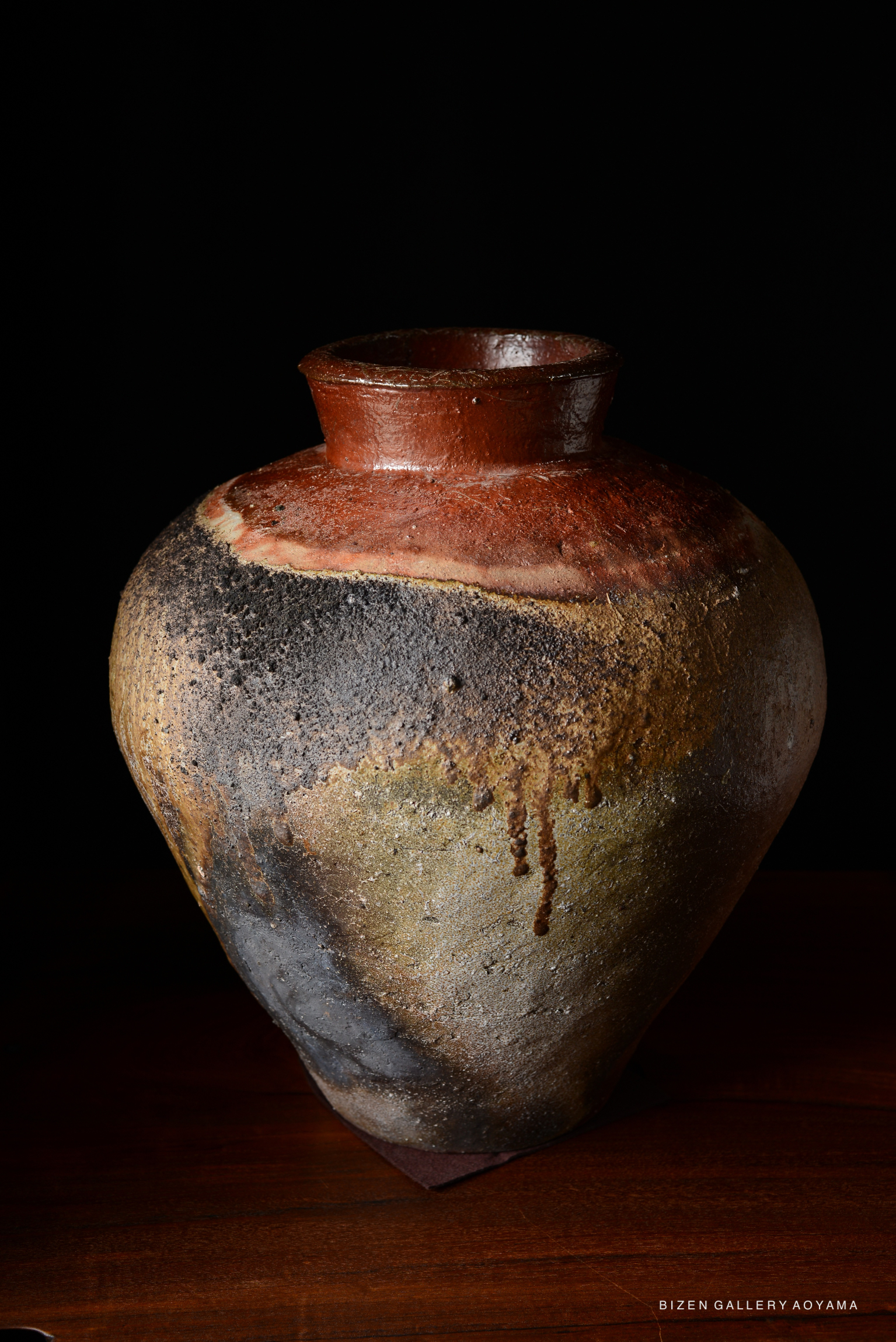 A Bizen tsubo, a traditional Japanese pottery vase, featuring an asymmetrical shape with a broad belly and narrow neck, showcasing a mix of earthy tones with natural texture and drips from the glaze, set against a black background.
