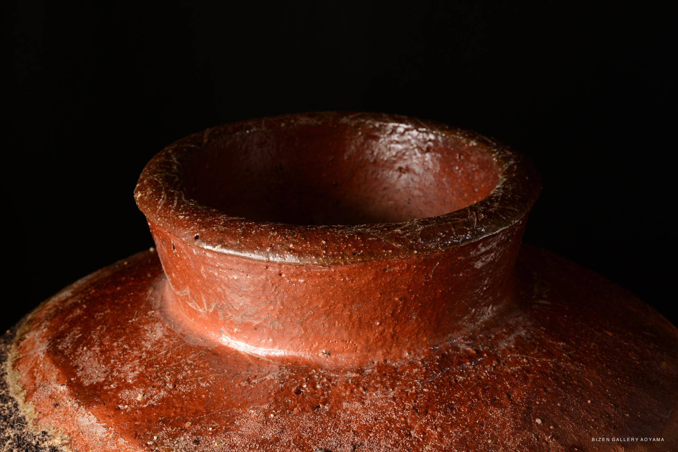 Close-up view of the rim of a Bizen pottery vessel, showcasing its textured surface and earthy reddish-brown color against a dark background.