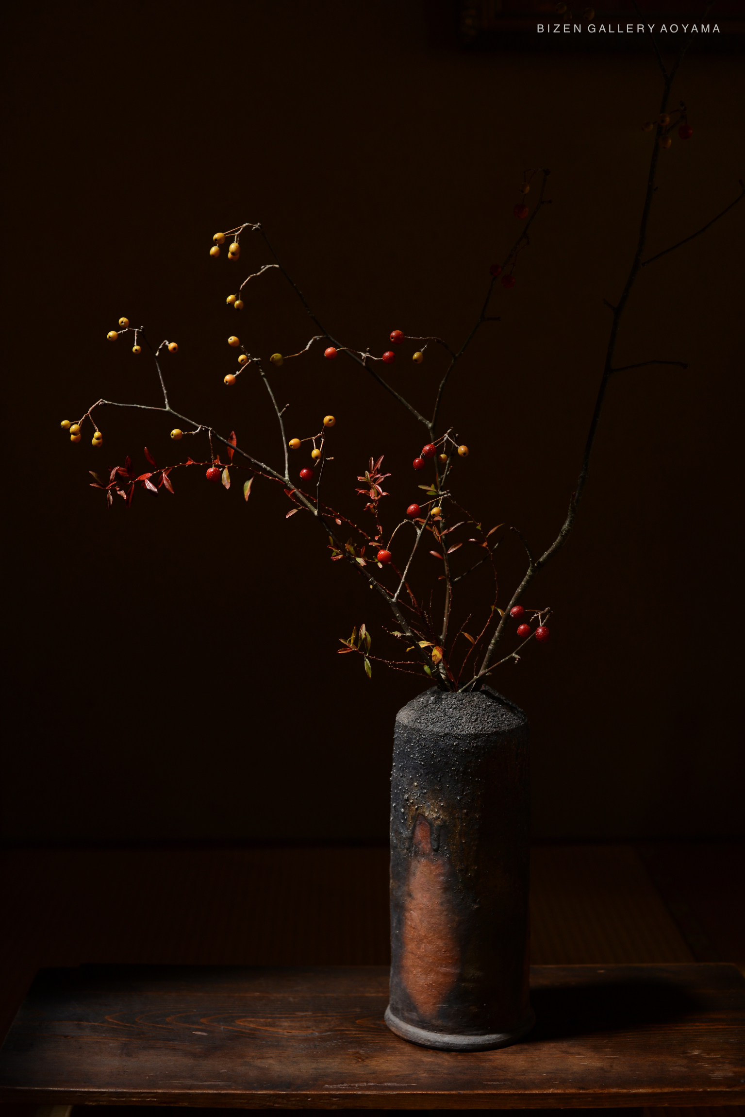A Bizen pottery vase with a textured surface, holding branches with small yellow and red berries, set against a dark background.