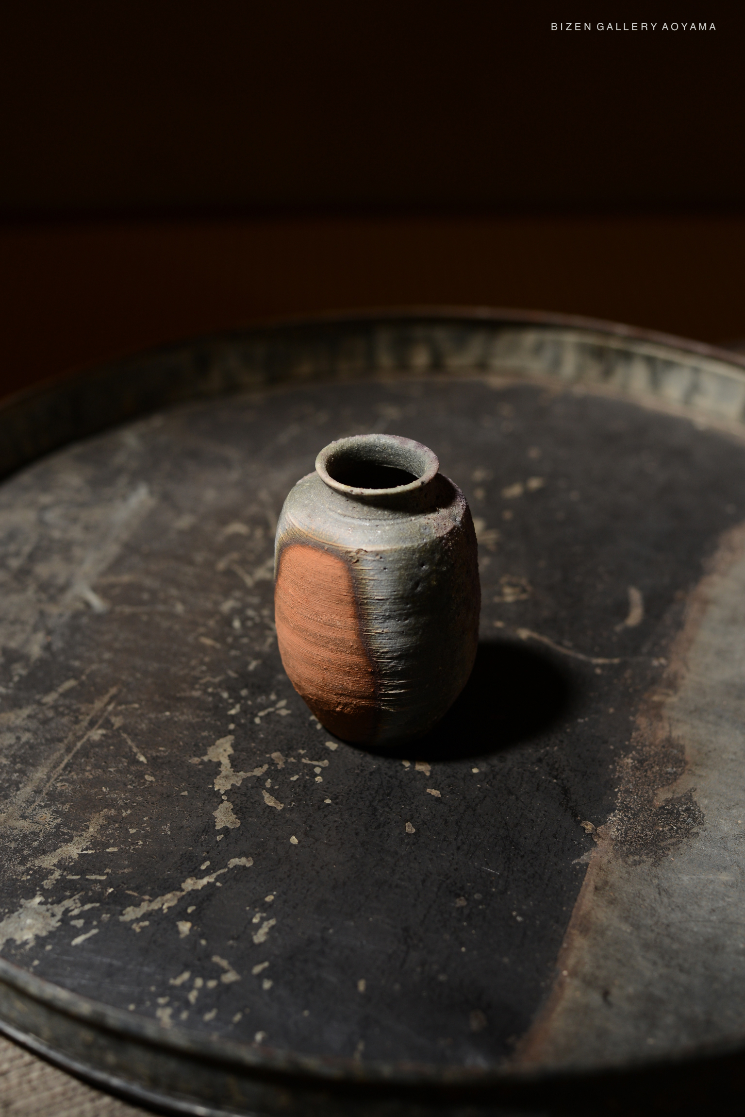 A close-up of a traditional Kotsubo (Chaire) ceramic vessel, featuring a textured surface with natural earthy tones, set on a dark metal tray.