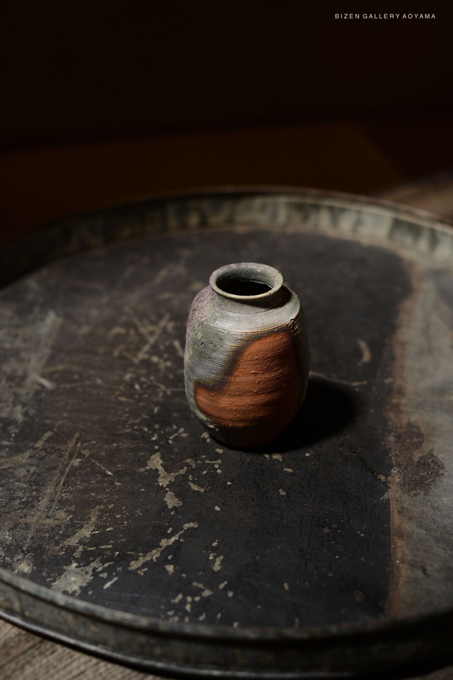 A Kotsubo (Chaire) pottery vessel placed on a textured dark tray, showcasing earthy colors and a smooth surface.
