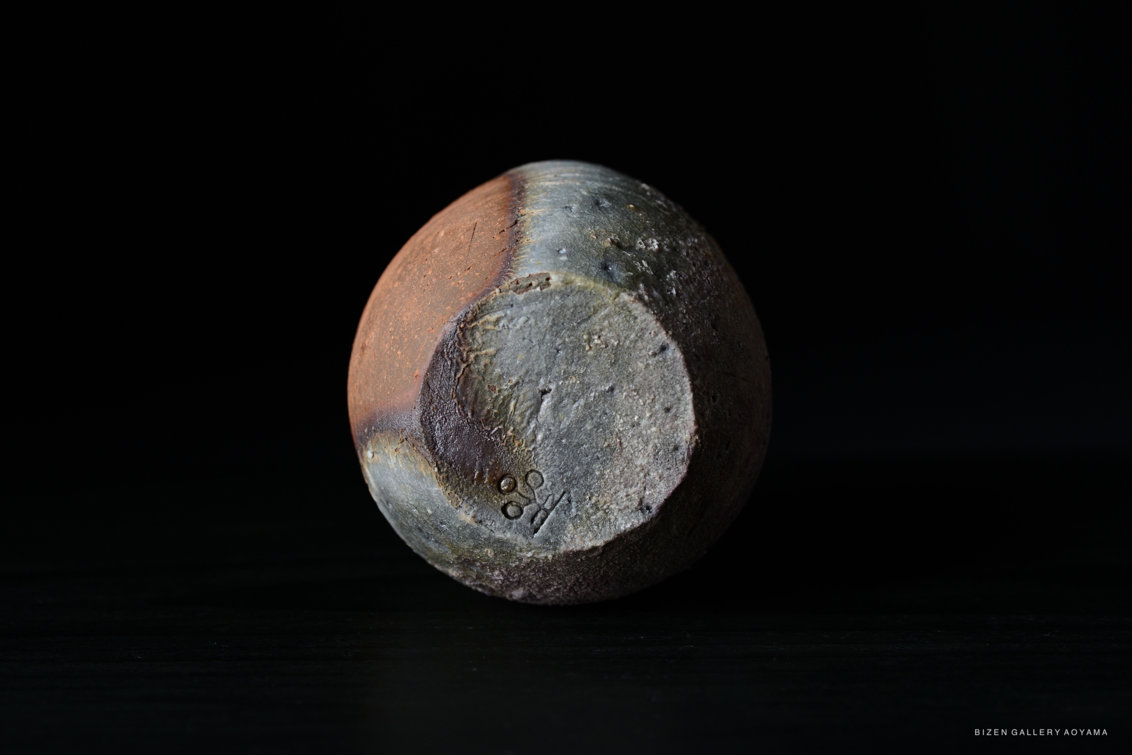 A close-up of a round Bizen pottery piece, showcasing its unique texture and color variations against a dark background.