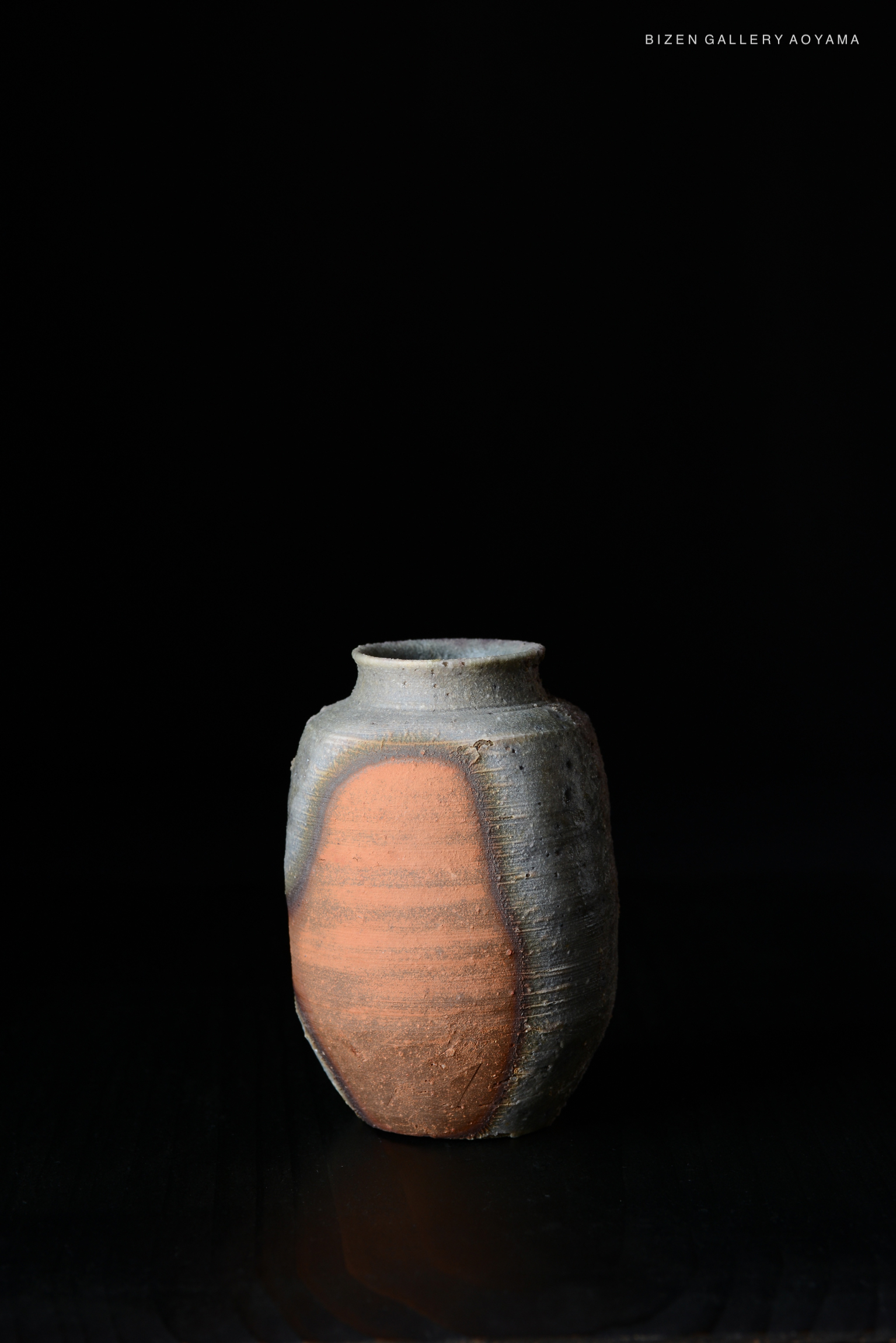 A small, round Bizen ceramic vase with a gray surface and a prominent orange-brown section, set against a dark background.