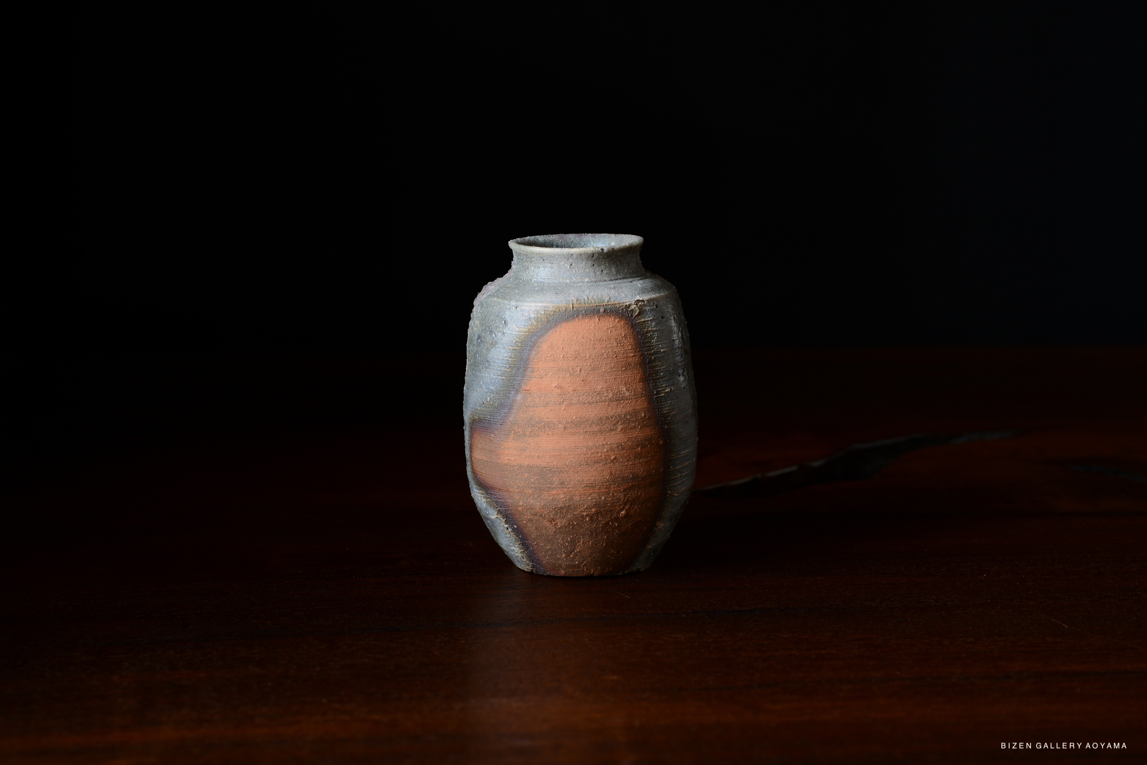 A close-up image of a traditional Kotsubo (Chaire) ceramic tea container with a textured gray and reddish-brown surface, set against a dark background.