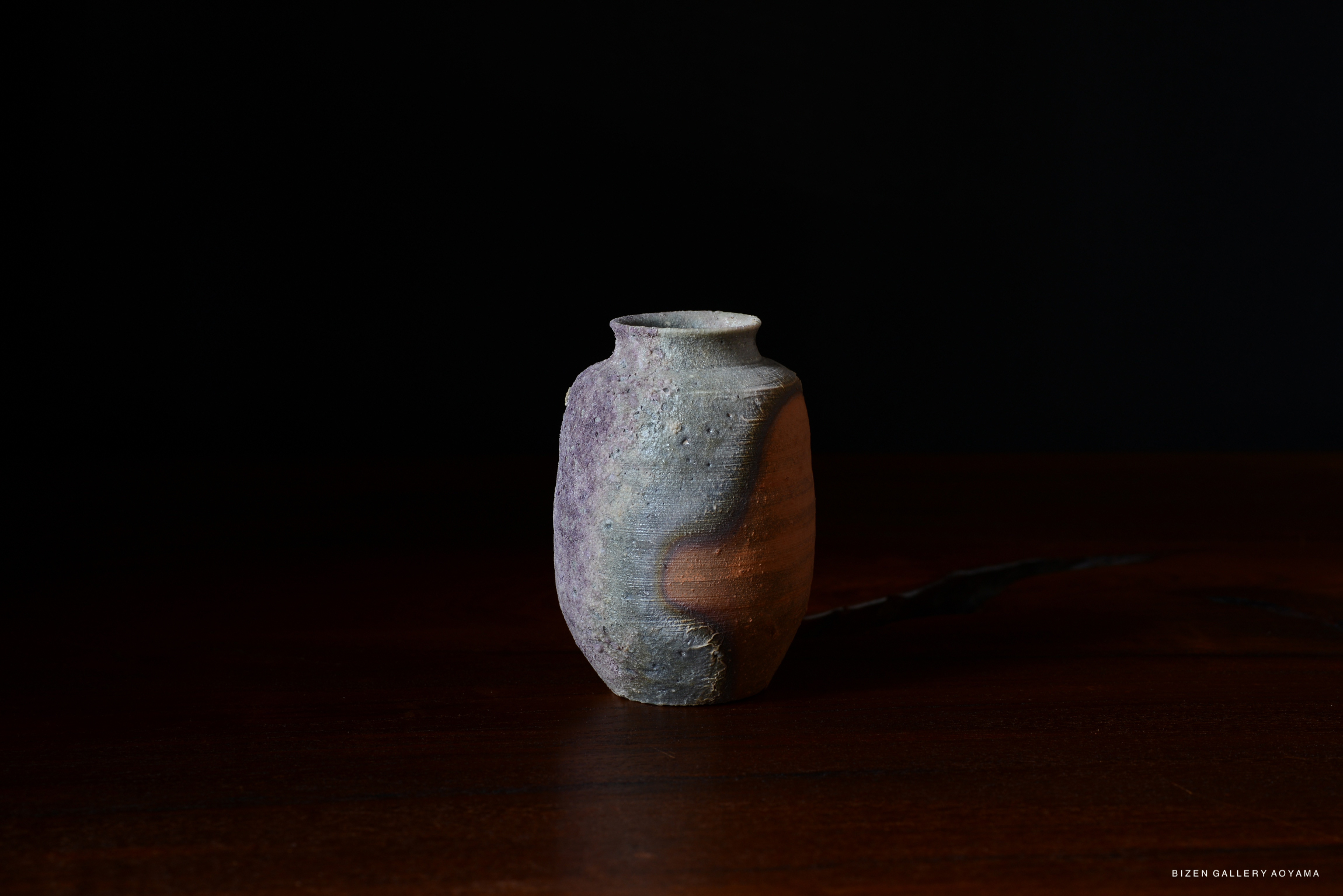 A small, rustic ceramic vase with a round body and a narrow neck, featuring a blend of earthy tones and textures, displayed on a dark wooden surface against a black background.