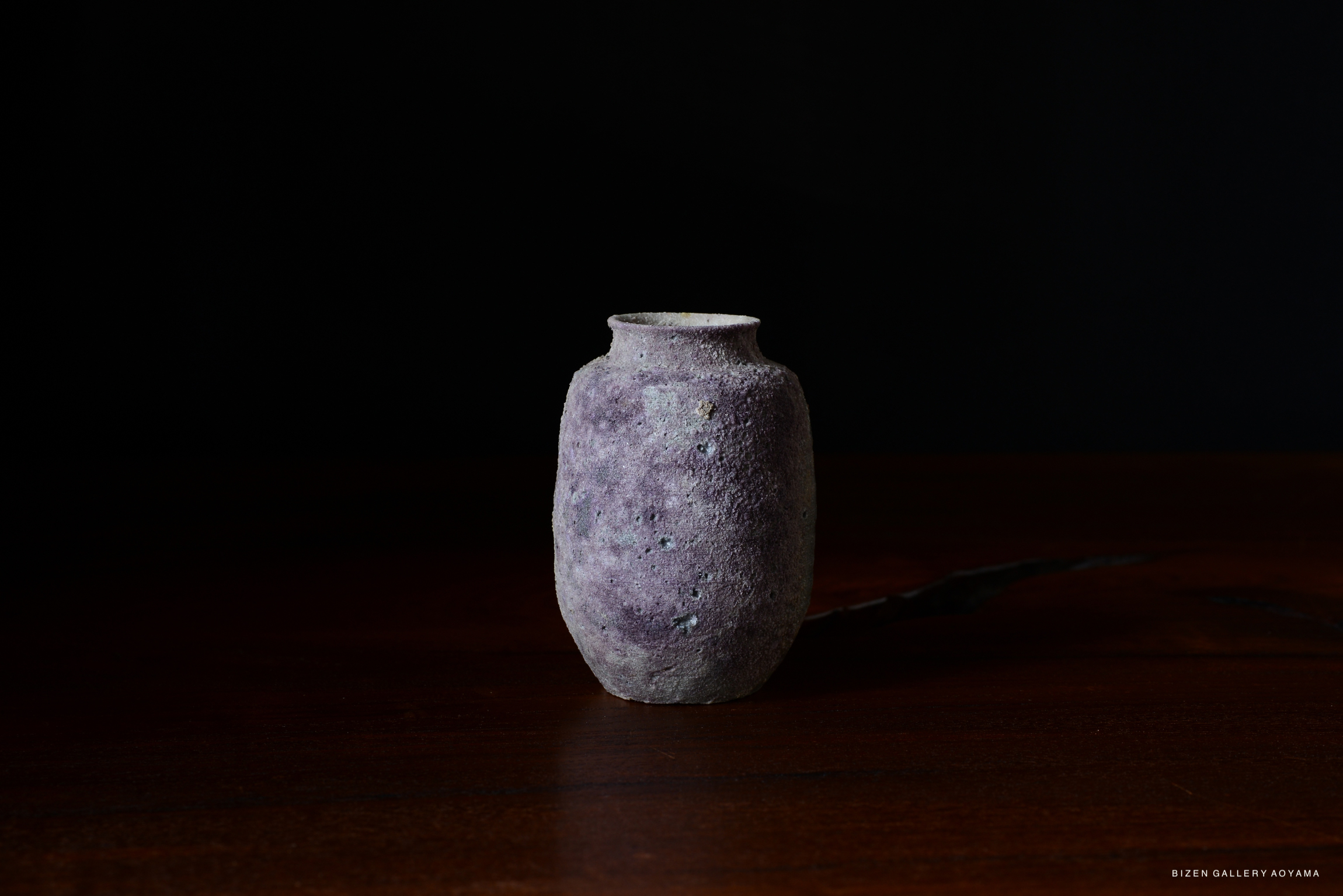 A rustic pottery vessel with a textured surface, featuring a muted purple color, placed on a wooden surface against a dark background.