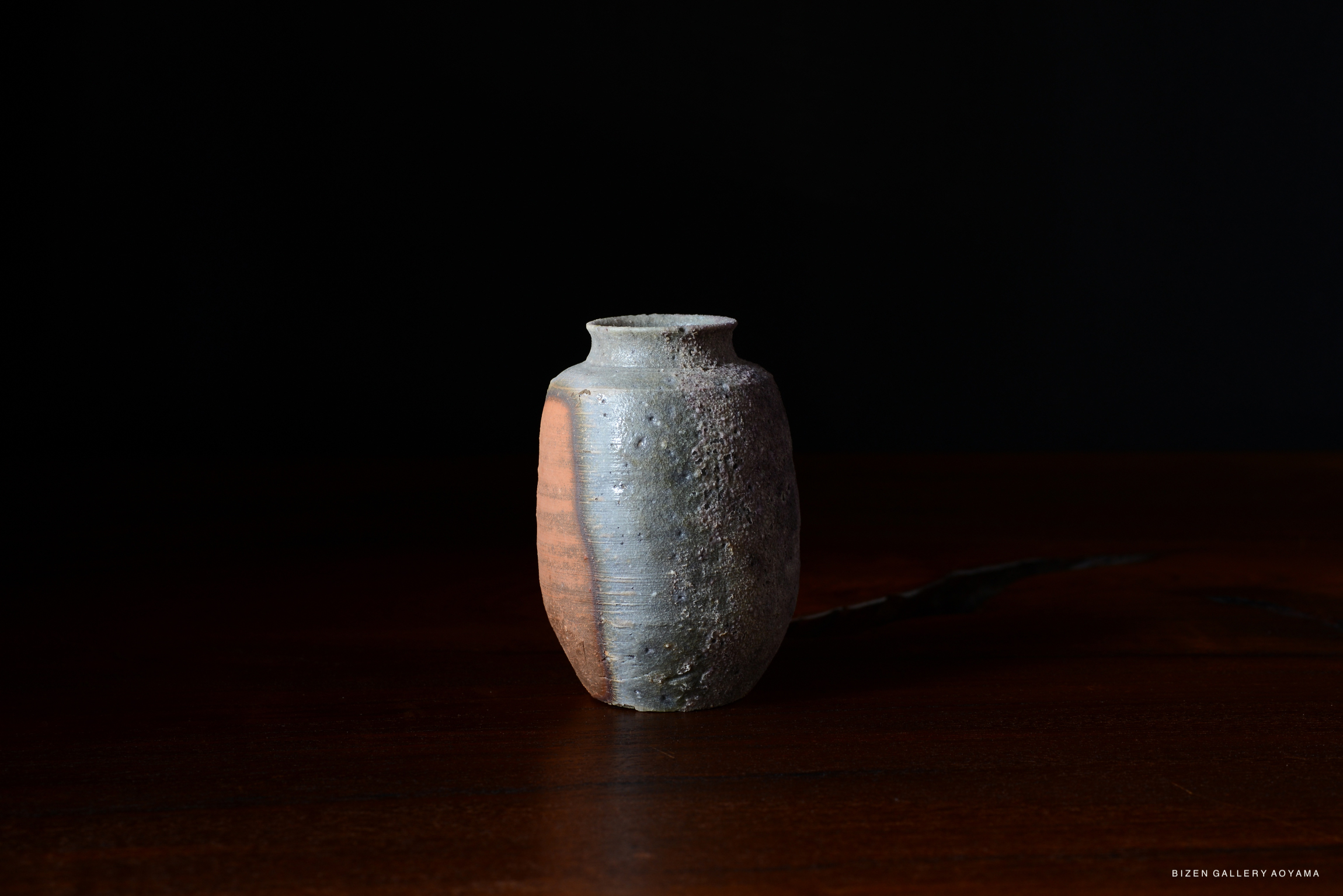 A small, textured ceramic vase with a rounded body and a narrow neck, featuring a mix of earthy colors, resting on a wooden surface against a dark background.