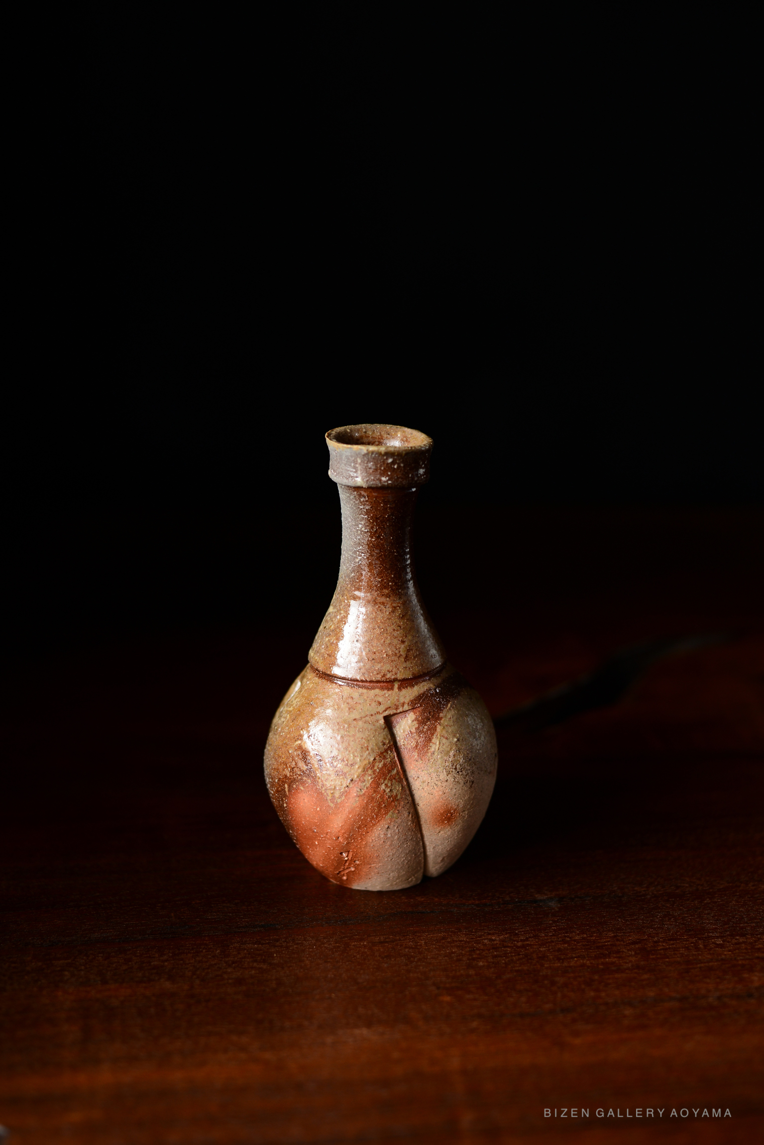 A Bizen tokkuri, a traditional Japanese ceramic sake bottle, featuring a distinctive round body and narrow neck, displayed on a wooden surface against a dark background.
