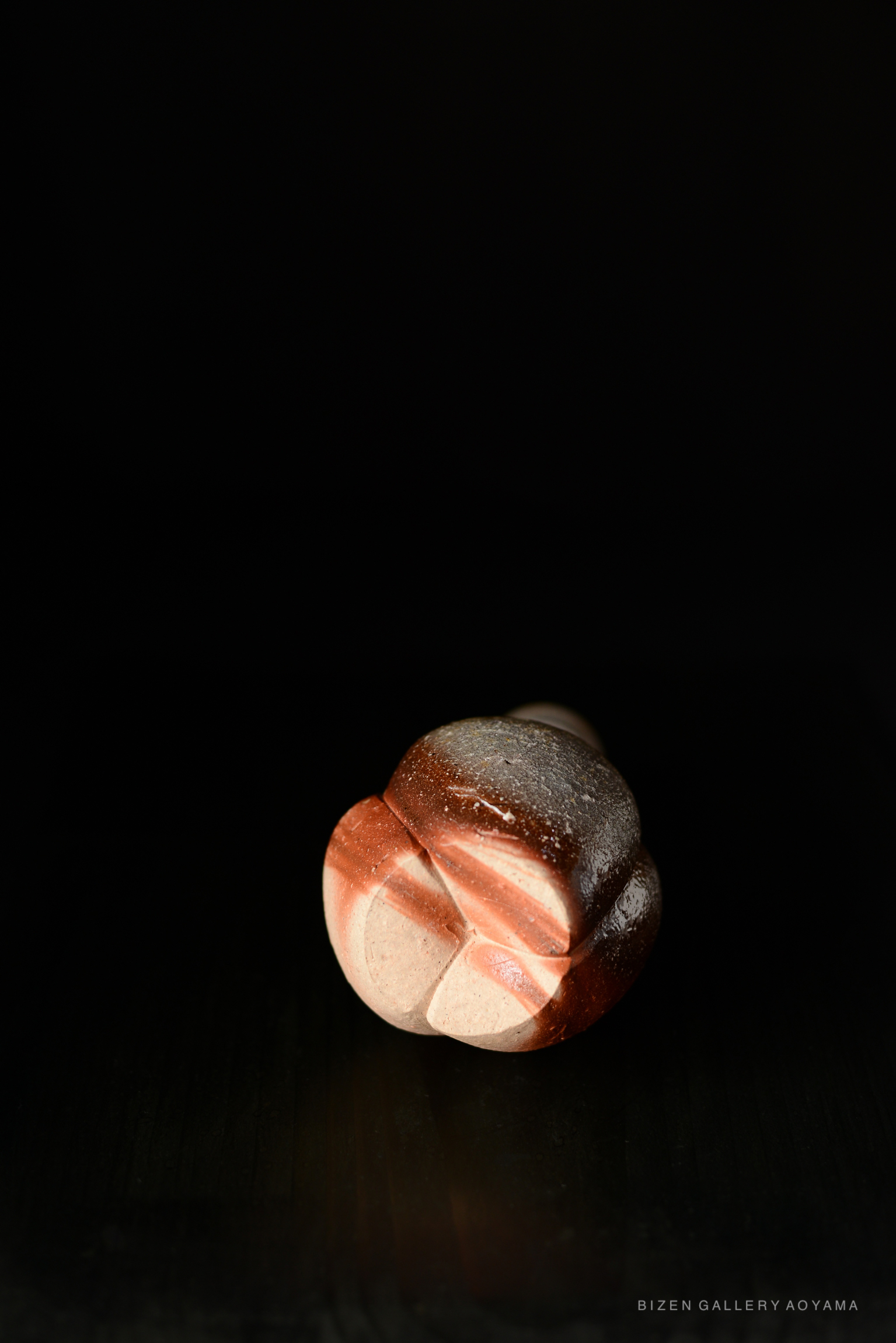 A Bizen Tokkuri with a unique shape, featuring a blend of earthy tones and textures, displayed against a dark background.