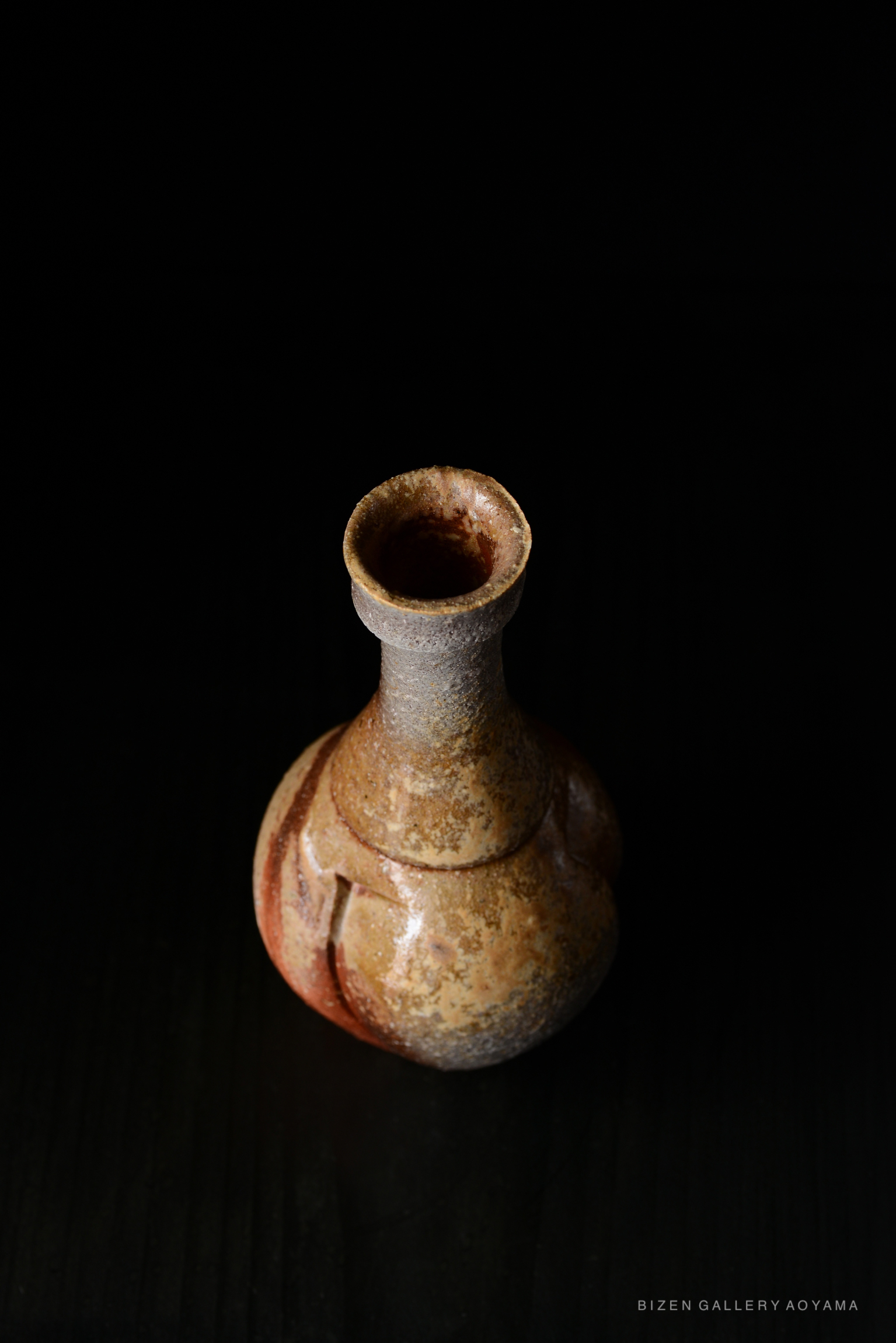 A Bizen pottery tokkuri with a rounded body and a narrow neck, featuring earthy tones and a textured surface, set against a black background.