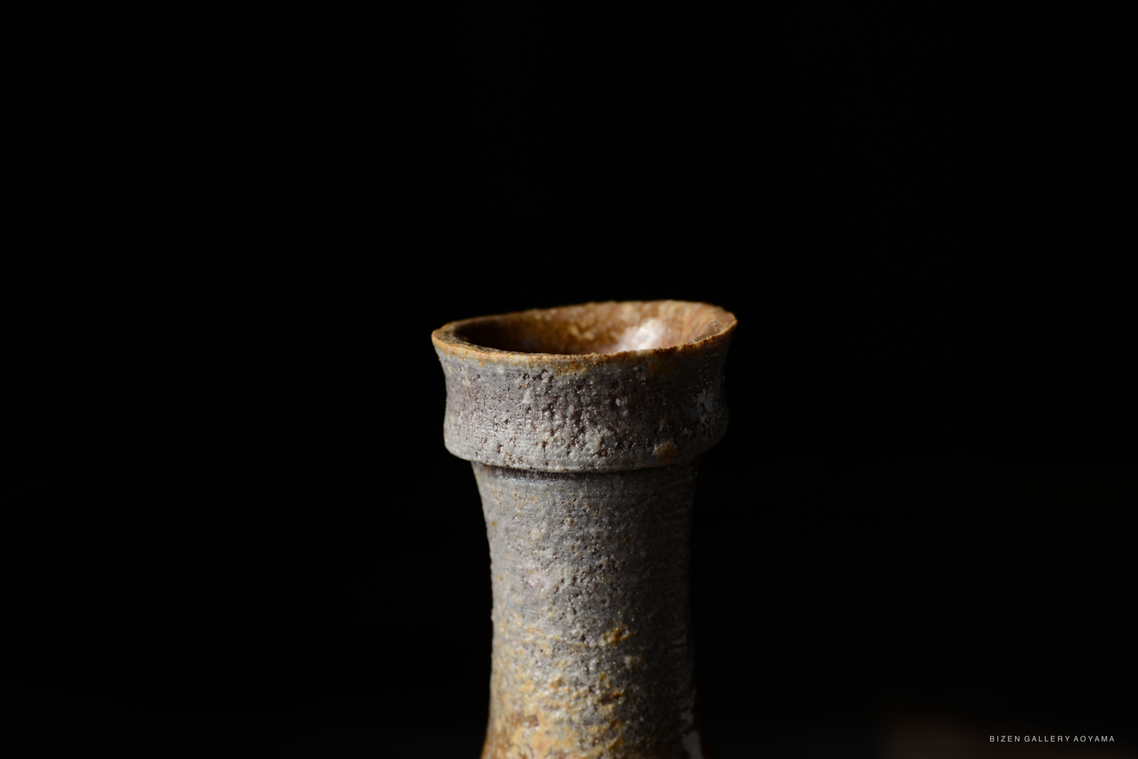 Close-up of a Bizen tokkuri (sake vessel) with a textured surface and a slightly flared rim against a dark background.