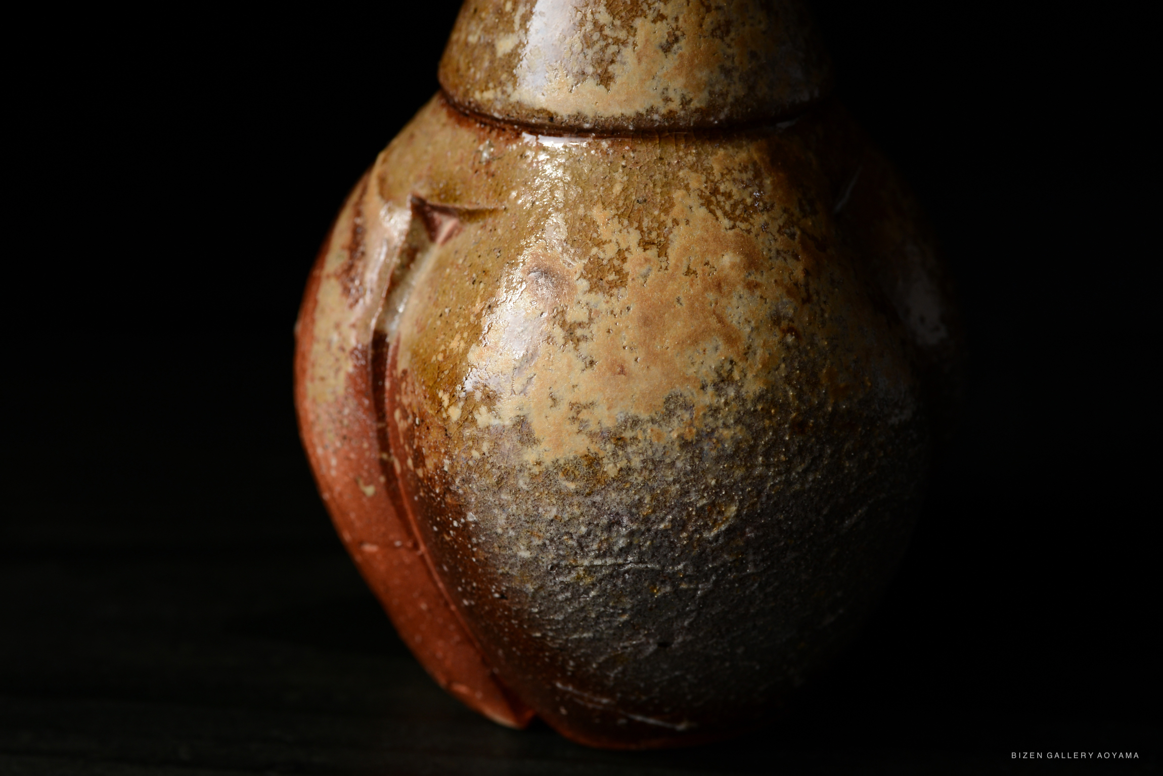 A close-up of a Bizen tokkuri, a traditional Japanese sake vessel, showcasing its unique textured surface and earthy colors against a dark background.