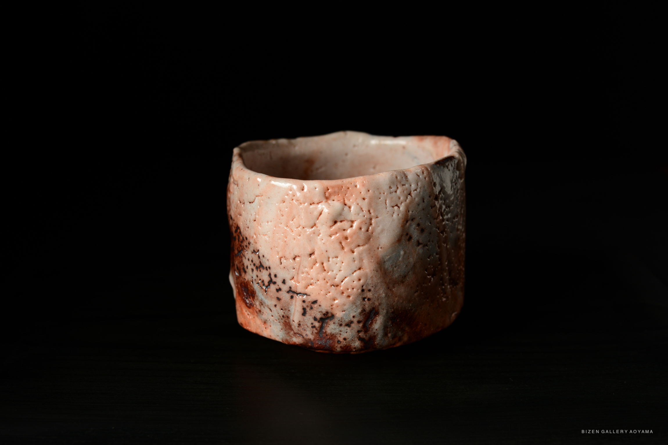 A close-up of a Shino Chawan tea bowl, showcasing its textured, earthy surface with a blend of pink and brown hues, set against a black background.