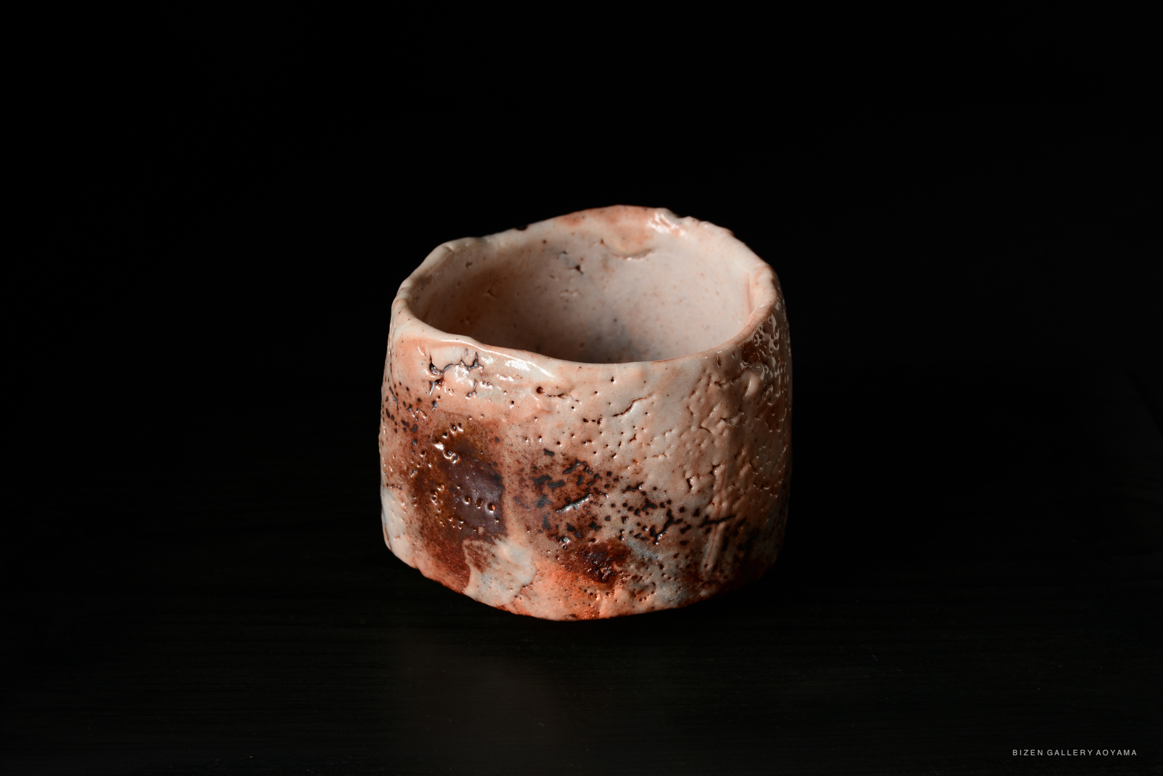 A unique Shino Chawan (tea bowl) with a textured surface and warm colors, displayed on a dark background, showcasing the artistry of Masayuki Higuchi.
