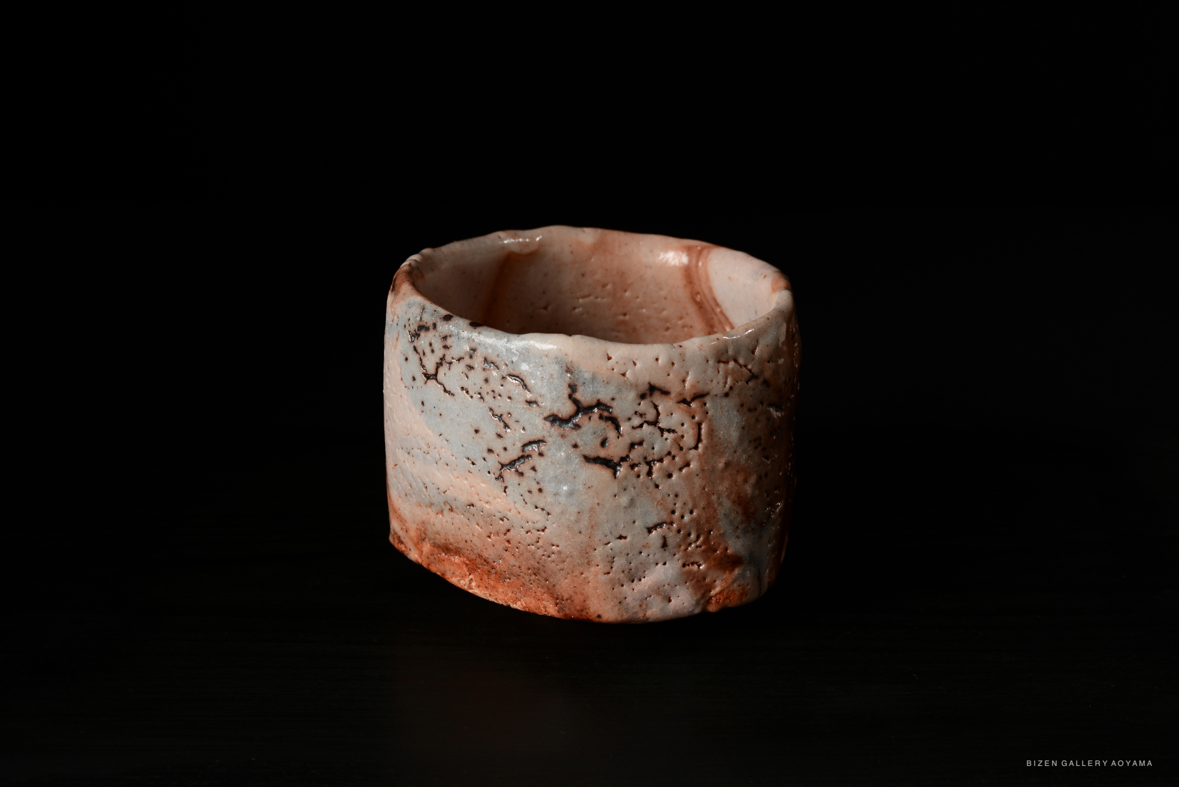 A close-up view of a Shino Chawan tea bowl featuring a textured surface with a mix of beige and reddish tones, set against a black backdrop.
