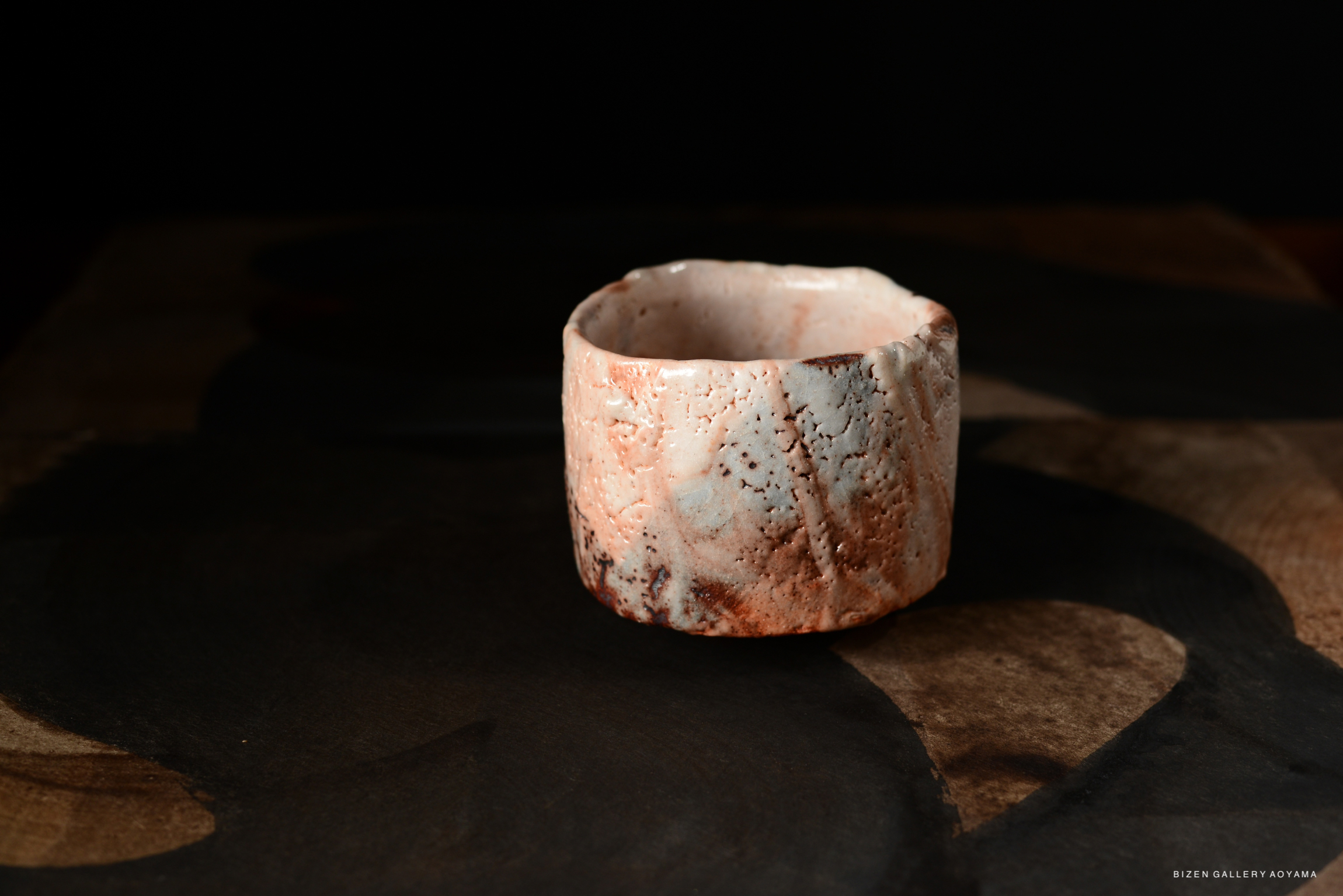 A close-up image of a ceramic Shino chawan (tea bowl), displaying a rustic texture with subtle earthy colors, placed on a dark wooden surface.