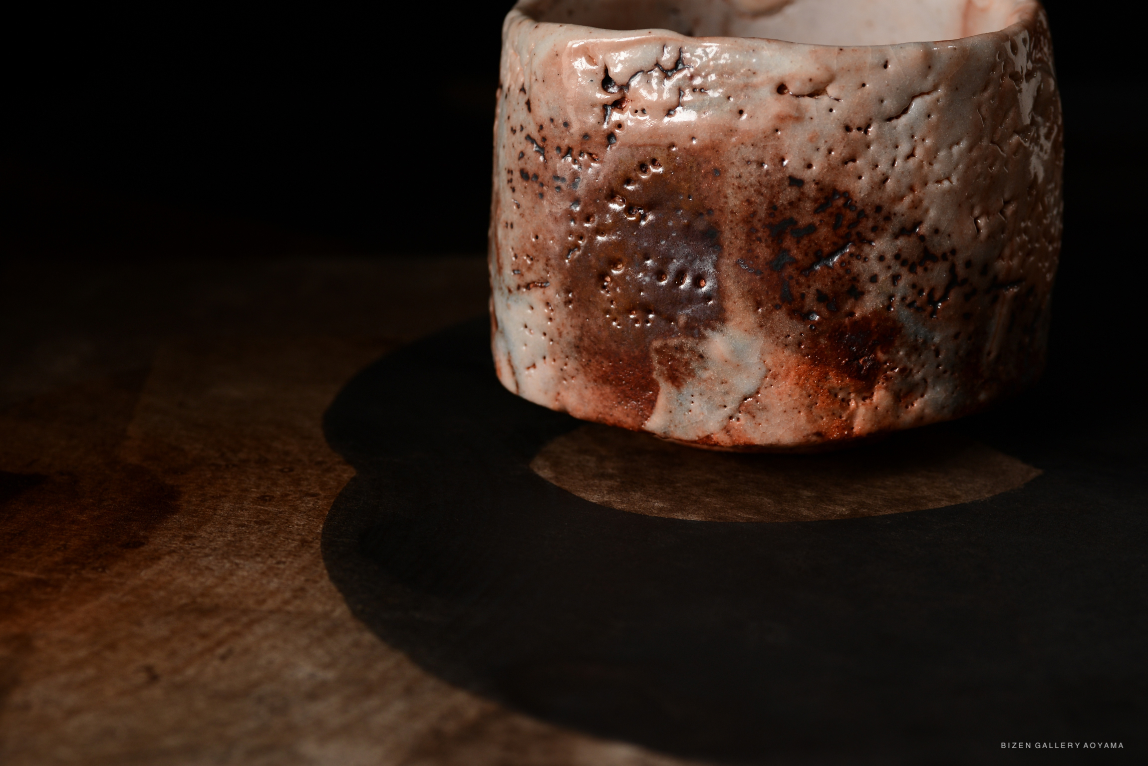 A close-up view of a Shino Chawan tea bowl showcasing a textured surface with a blend of earthy tones and a glossy finish, set against a dark background.